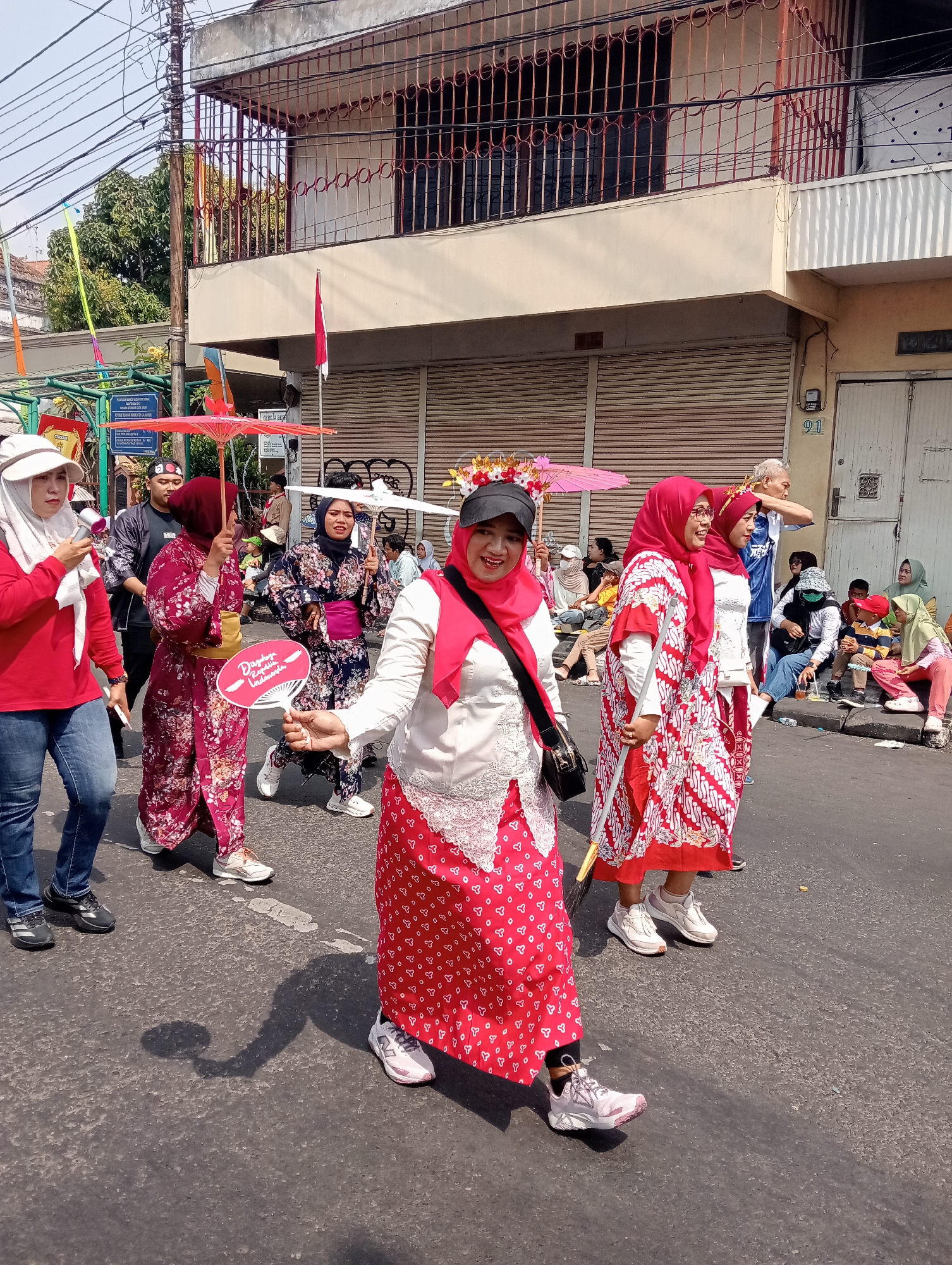 In Bintoro, Indonesia, a group of women are participating in a parade. The women are dressed in traditional attire, including colorful kimonos and headscarves. They are walking down a street lined with buildings and power lines. Some of the women are carrying umbrellas and fans, while others are holding small flags. In the background, a crowd of spectators can be seen sitting on the side of the road, watching the parade. The scene appears to be festive, with a sense of community and celebration.