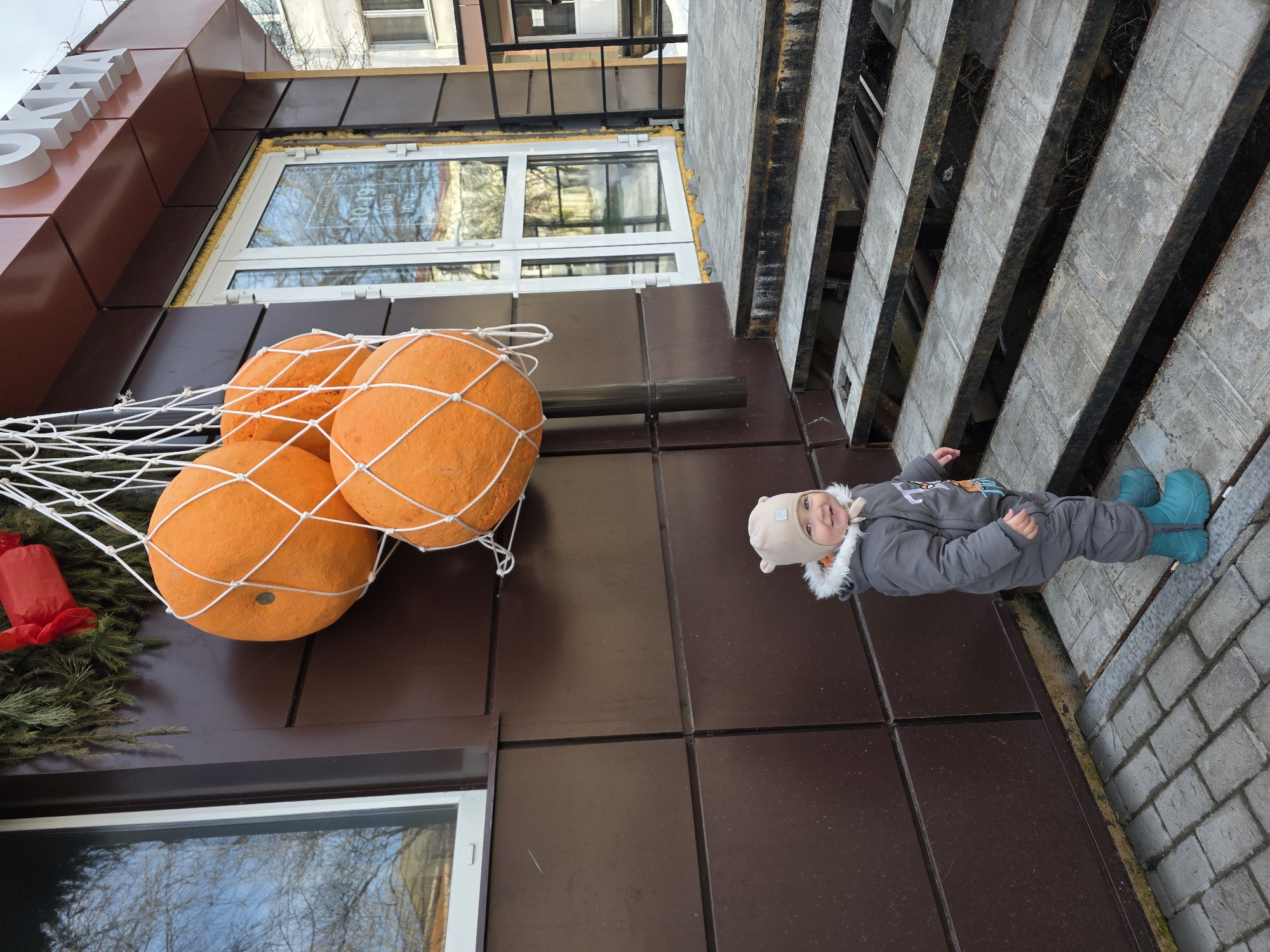 An outdoor scene in Chaikovsky, Russia, features a toddler standing on a paver-stone sidewalk against a brown, horizontally paneled building facade. Above the child, decorative elements include an artificial green pine wreath with a red accent, from which three large, orange, spherical objects resembling oversized oranges are suspended within a white mesh net. To the child's right, a set of worn metal and concrete stairs ascends to a white-framed glass double door. The door displays text including operating hours (e.g., "10-19") and partial Cyrillic signage indicating "ОКНА" (Windows). The child, dressed in a grey fur-hooded winter jumpsuit, a beige beanie with ear-like protrusions, and teal boots, stands facing slightly towards the viewer, smiling. A building window to the left reflects bare tree branches under an overcast sky, indicating an outdoor urban environment.