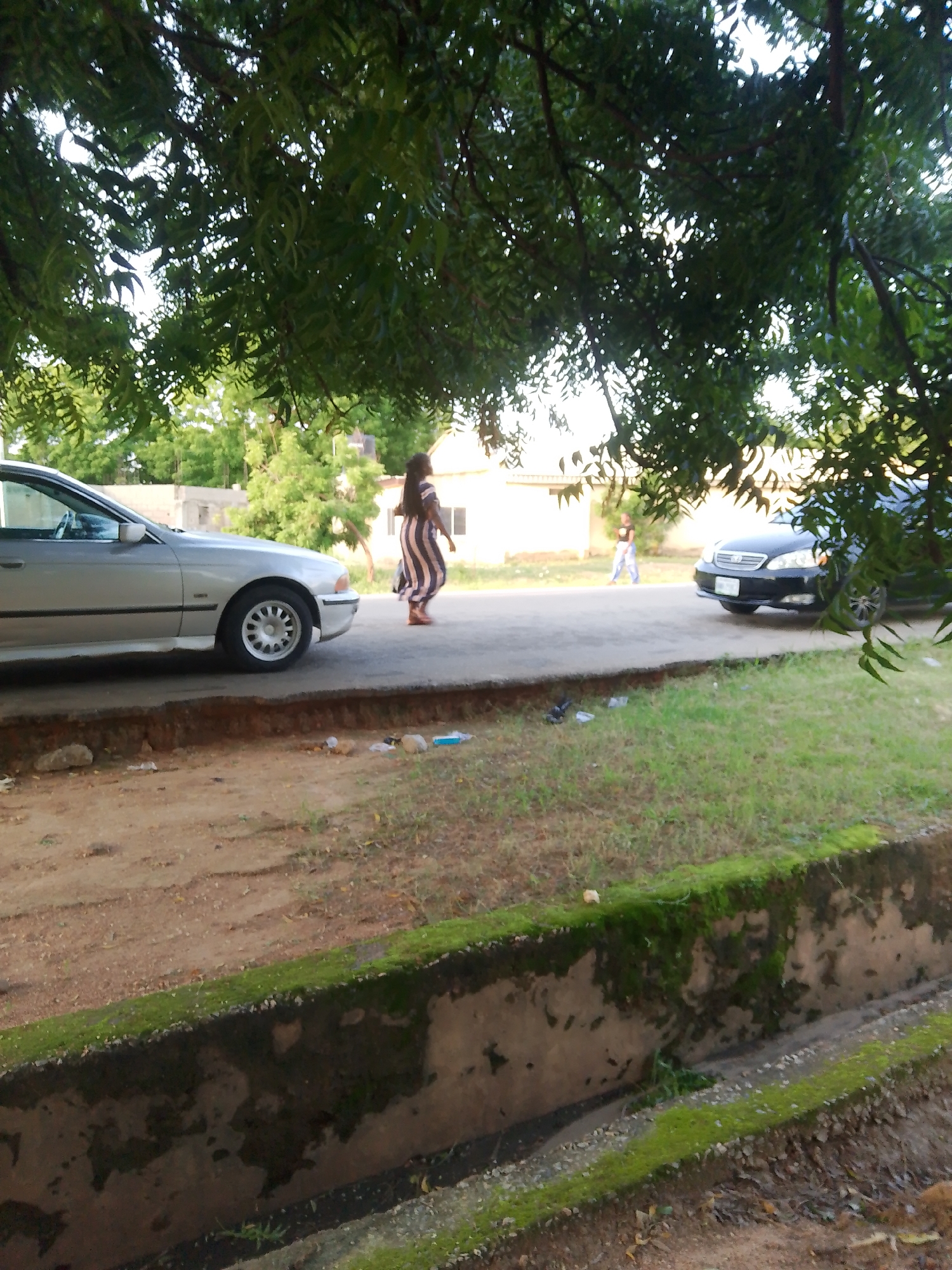 A silver sedan is parked on the side of a road in Jalingo, Nigeria. The car is facing left. A woman wearing a striped dress is walking on the road behind the car, facing away from the camera. A black sedan is parked on the other side of the road, facing right. There are trees and buildings in the background. The foreground shows a ditch with moss-covered concrete walls and dirt.