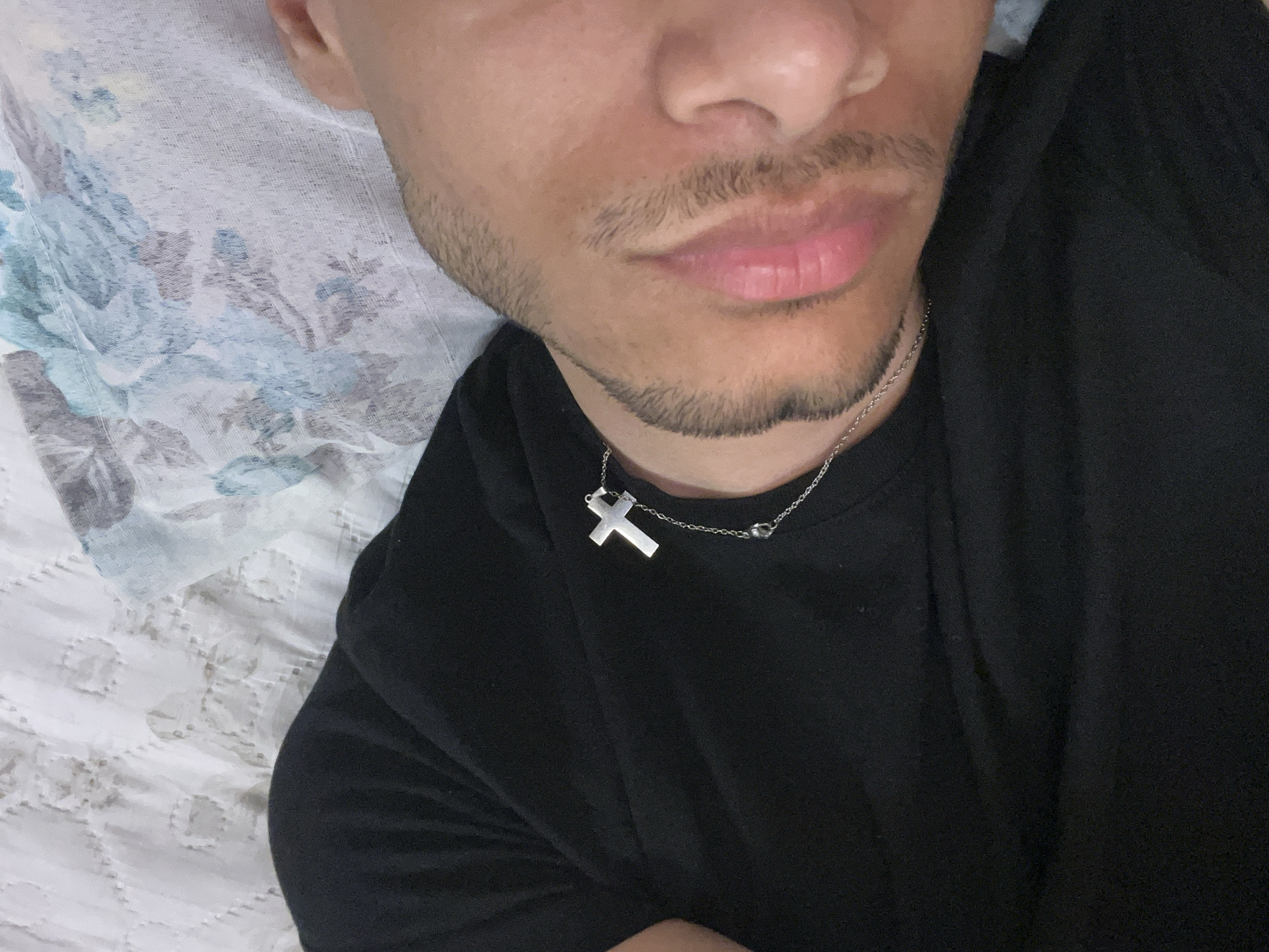 A close-up, high-angle shot captures the lower half of a person's face and their upper torso, likely a selfie taken while reclining. The person, appearing male, has short facial hair, including a mustache and a goatee. They are wearing a plain black t-shirt or top and a thin silver-colored chain with a silver cross pendant around their neck. The subject is resting on a surface, likely a bed or sofa, indicated by the bedding. To the left, a light-colored pillow or blanket features a muted floral pattern with shades of blue, teal, and brown. Below that, on the right side of the frame, is another white, textured blanket or sheet with a subtle, circular quilted pattern. The lighting is soft and ambient, suggesting an indoor setting. No specific activity is depicted other than resting, and no strong emotions are conveyed. There is no visible text.
