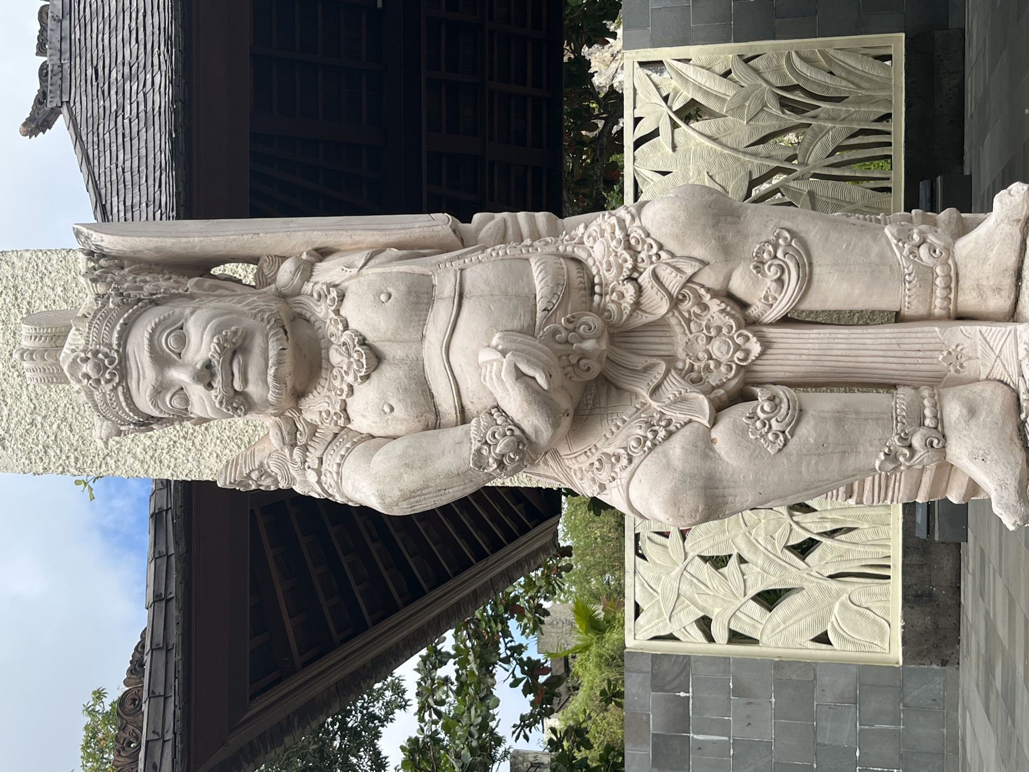 The image shows a stone statue of a Balinese guardian in front of a building with traditional Balinese architecture, likely located in Kuta Selatan, Indonesia. The statue is a stout, male figure with intricate carvings, wearing a headdress and ornate jewelry. It stands in what appears to be an outdoor courtyard or walkway. Behind the statue, a weathered wooden roof with intricate carvings is visible, supported by columns. A decorative screen with a leafy pattern is also present in the background, along with gray stone walls and tiled flooring. The sky is overcast with patches of blue.
