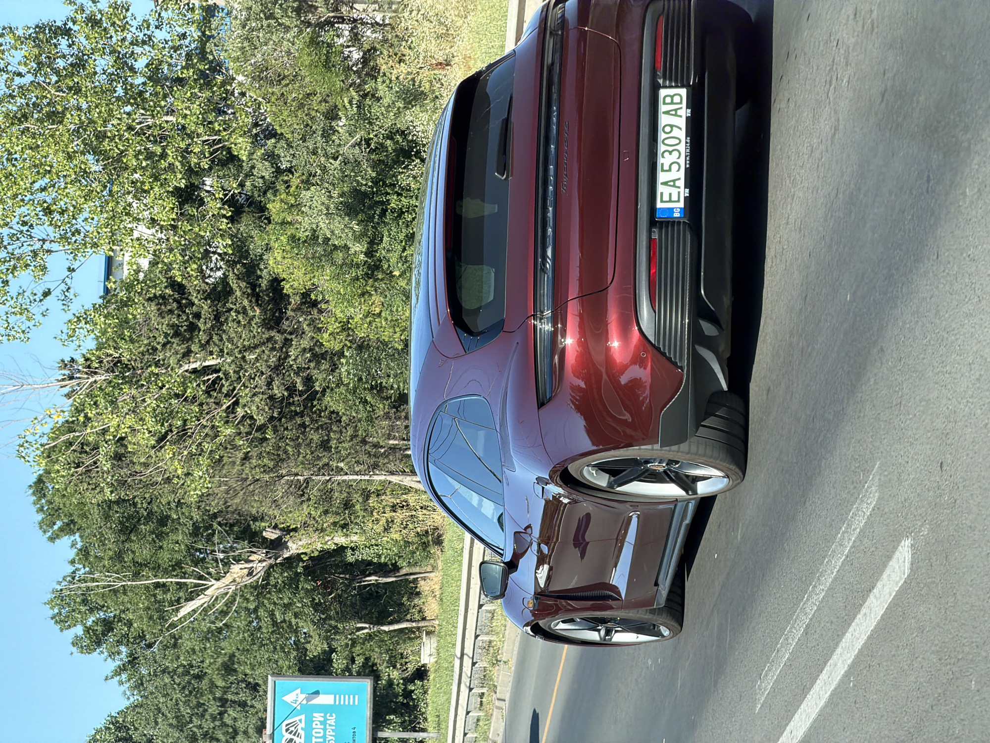 The media file shows the rear-left side of a dark red Porsche Taycan GTS parked on a paved road. The car has a sleek design with a prominent spoiler and distinctive taillights. The license plate, EA 5309 AB, is clearly visible and indicates Bulgaria as the country of origin (BG). A website, www.TR24.de, is also printed on the license plate frame.
The background consists of lush green trees and a clear blue sky, suggesting it is daytime with fair weather. On the left side of the frame, a blue sign with white text is partially visible. The text on the sign appears to be in Bulgarian, with "ТОРИ" and "БУРГАС" being legible, indicating Burgas, Bulgaria, as a potential location. The overall scene is one of a car on a road, likely in a somewhat urban or suburban area given the presence of the road markings and signage. There are no people visible in the image. The sun appears to be casting reflections on the car's polished surface, highlighting its curves and the metallic sheen of the paint.