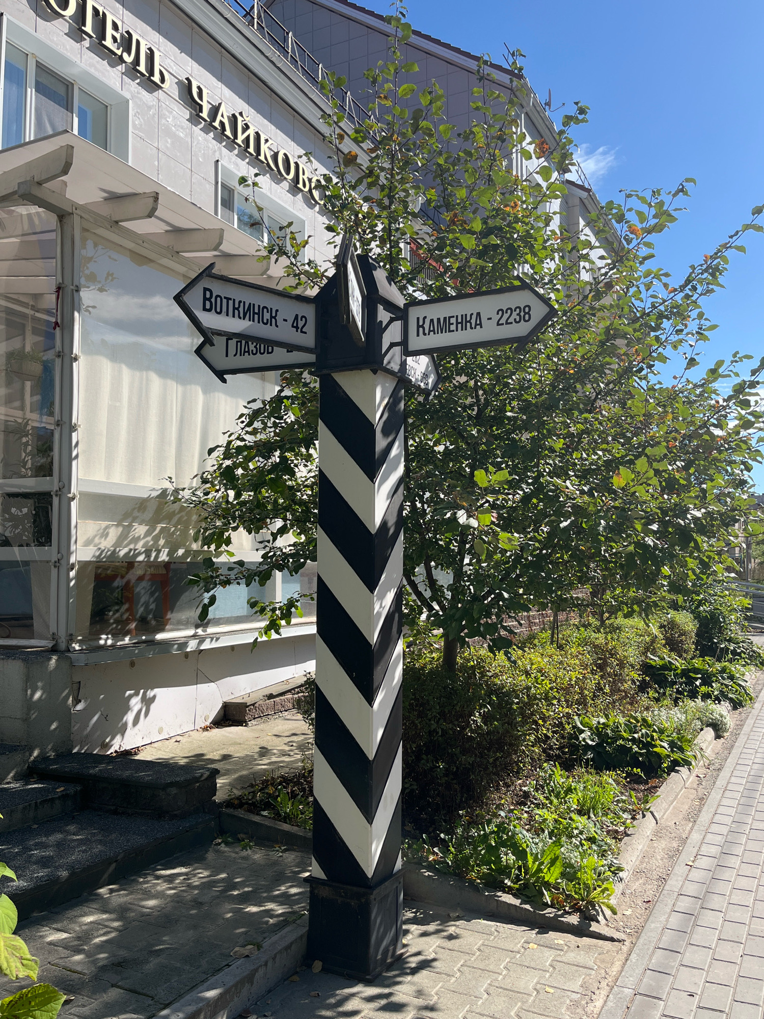 A black and white striped signpost with three directional signs stands on a sidewalk in front of a building. The signpost is in the foreground, to the right of center. The signs point left and right. The top left sign reads "ВОТКИНСК - 42" (Votkinsk - 42) and points left. Below that, another sign points left and reads "ГЛАЗОВ" (Glazov). The top right sign reads "КАМЕНКА - 2238" (Kamenka - 2238) and points right. The building behind the signpost is light-colored with windows. The top of the building has large gold letters that read "ОТЕЛЬ ЧАЙКОВСКО" (Hotel Chaykovsky). A tree with green leaves is to the right of the signpost, obscuring part of the building. The sidewalk is paved with gray rectangular stones, and a raised garden bed with green plants is between the sidewalk and the building. The sky is blue. The scene appears to be in the city of Chaikovsky, Russia, as indicated by the hotel's name. There are no people visible in the image.