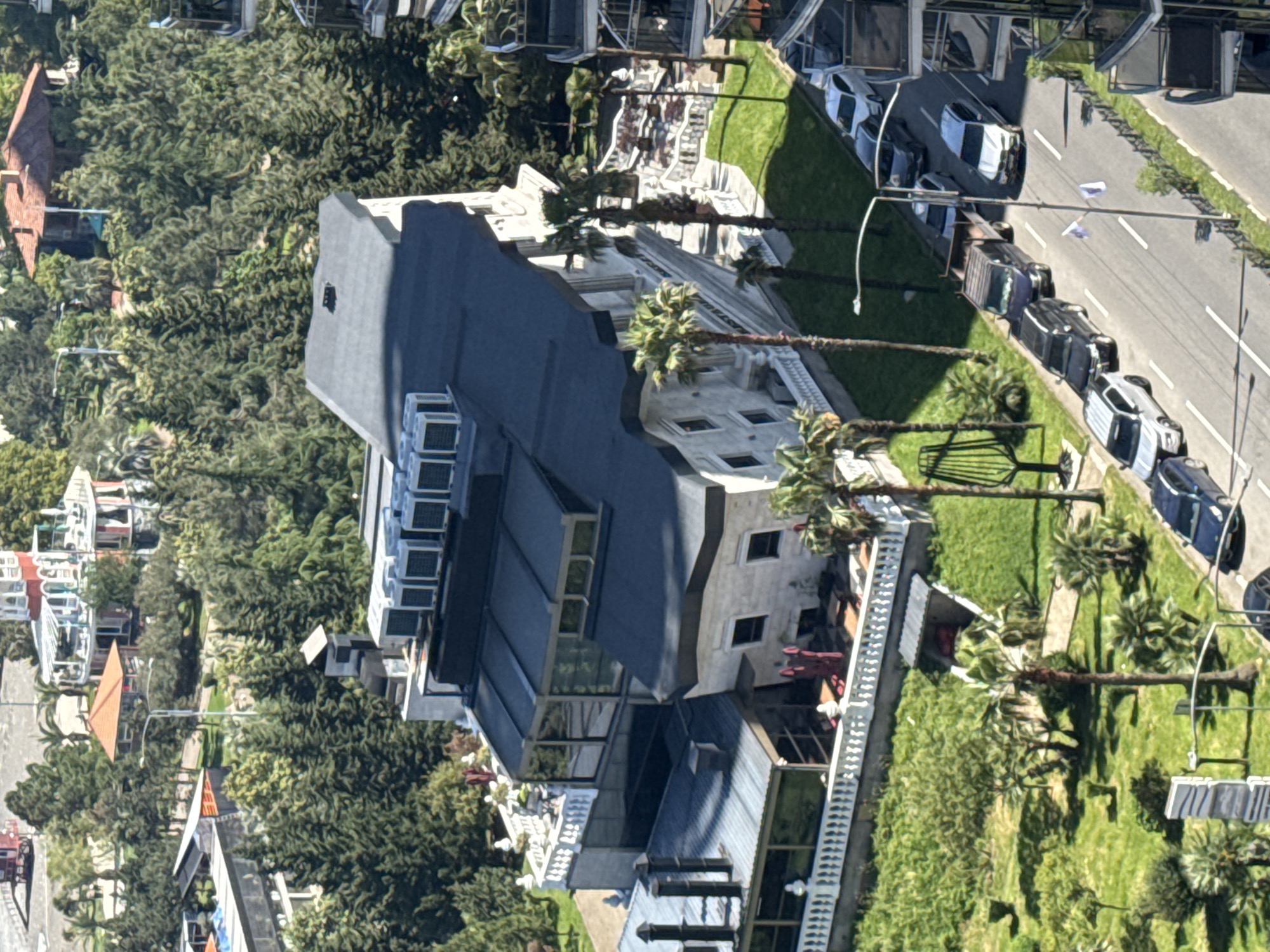 An eye-level aerial shot depicts a tall, modern building with a tiered, slate-gray roof, located in Batumi, Georgia. The building is surrounded by lush greenery, including mature trees and manicured lawns interspersed with palm trees. The building's facade is a mix of white concrete and large windows, reflecting the sky. A row of sun loungers is visible near the building, suggesting a resort or hotel. Adjacent to the lawn, a road is lined with parked cars, some of which are white. A light pole with flags is positioned along the road. In the background, other buildings with red-tiled roofs are partially obscured by the trees. The overall environment suggests an urban resort setting. There are no visible people in the image.