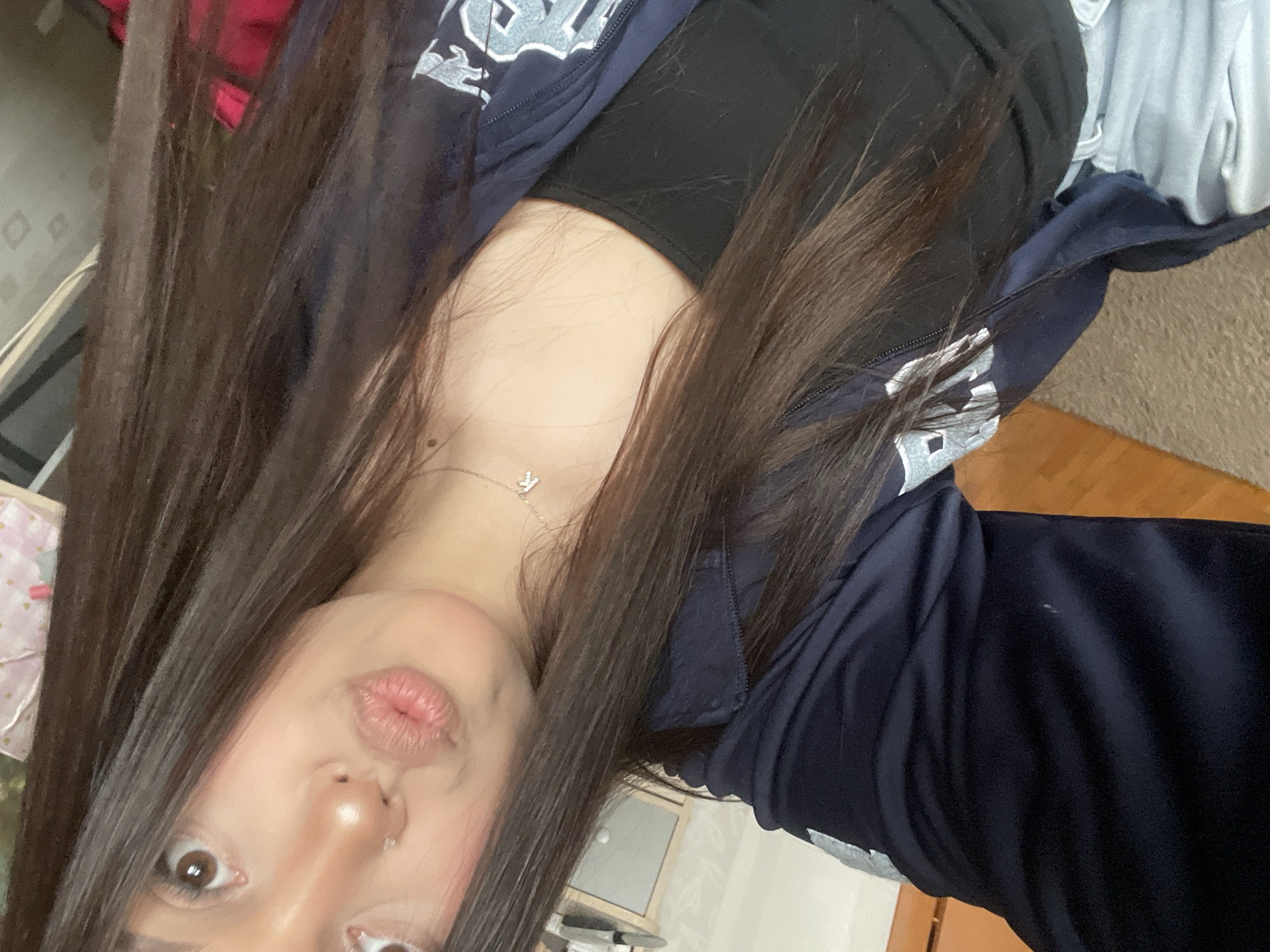 A young woman is captured in a close-up selfie, taken from a slightly high angle and rotated counter-clockwise, showing her from the chest up. She has long, dark brown hair that frames her face and flows across parts of the image. Her skin appears fair, and she has a nose piercing on her right nostril. Her lips are puckered in a "kissing" pose, and her brown eyes are wide open, looking directly at the camera with a playful expression.
She is wearing a dark navy blue athletic jacket with a zipper, open to reveal a black top or sports bra underneath. On the left side of her jacket, near her shoulder, there's a light blue or white stylized logo or letters, possibly "WS" or a similar design. Around her neck, she wears a delicate silver chain necklace with a small, sparkling pendant that resembles a stylized bird or the letter 'M' or 'W'.
The background is softly blurred but suggests an indoor setting, possibly a room in a home in Vsevolozhsk, Russia. Hints of a light-colored wall or furniture, a wooden floor or surface, and some light-colored fabric (perhaps clothing or a blanket) are visible. A splash of red color is also faintly discernible in the upper left corner. The lighting appears to be ambient indoor light. There is no clearly legible text other than the stylized logo on her jacket.