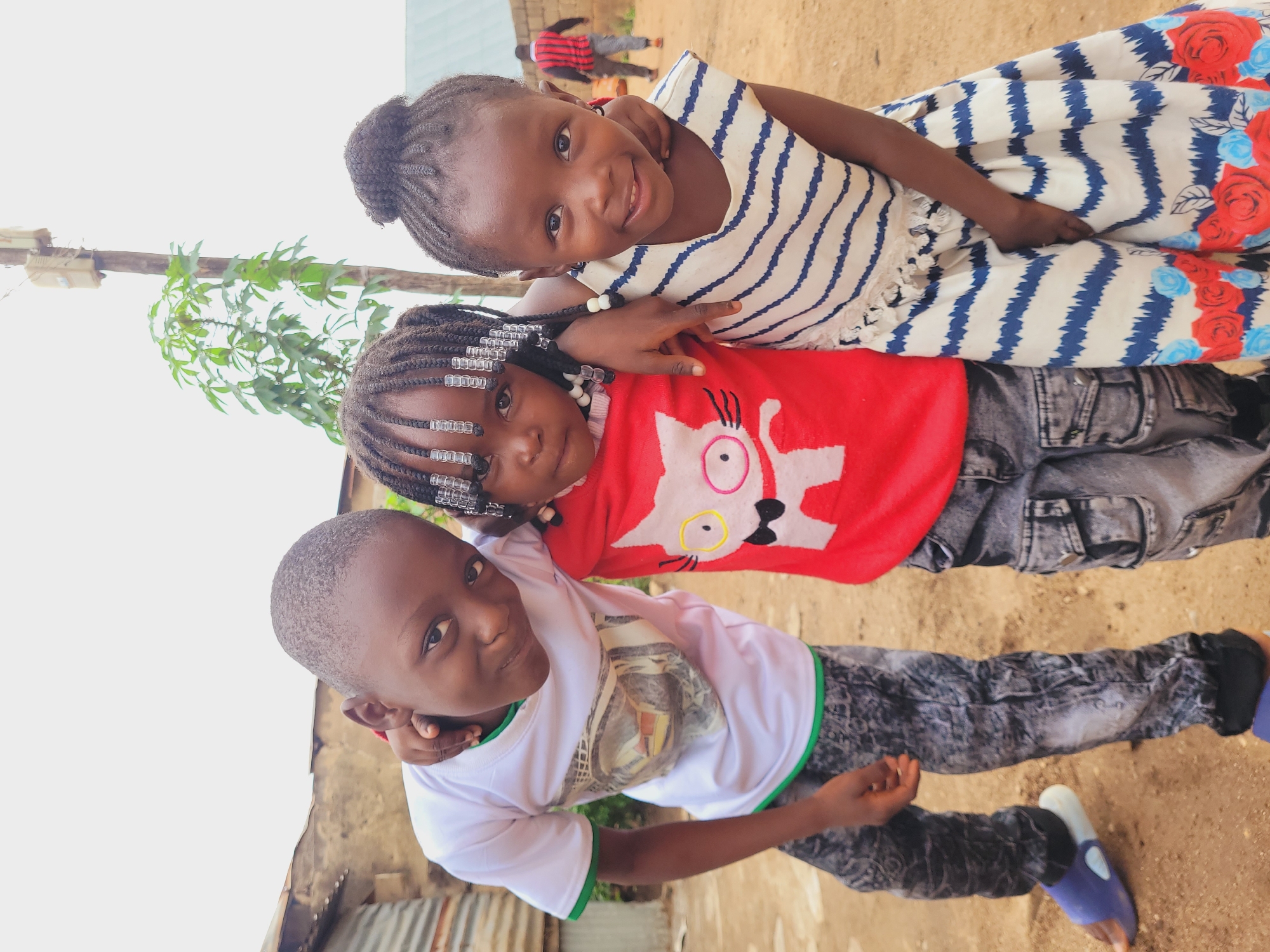 Three children are posing for a photograph. The tallest girl, wearing a white dress with blue stripes and red floral patterns, is in the background. A younger girl wearing a red shirt with a cat design is in the middle. A young boy wearing a white t-shirt with green trim is in the foreground, his arm around the younger girl. They are outside, on a dirt surface. In the background are glimpses of corrugated metal structures and a person in a red striped shirt. The location is Bukuru, Nigeria.