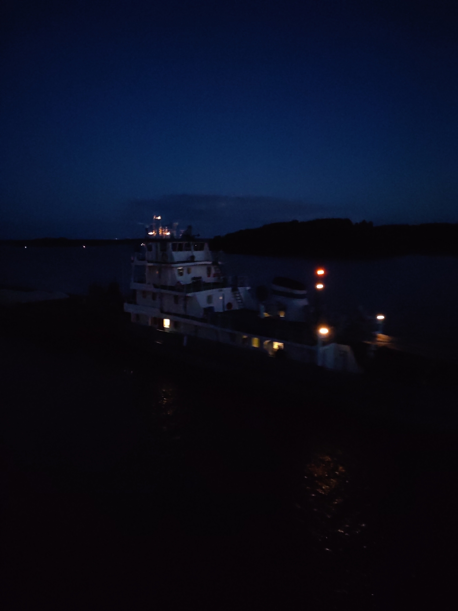 The image shows a large tugboat illuminated by internal and external lights in a dark body of water, presumably at night. The boat is the primary subject, displaying a multi-level superstructure with various light sources visible through windows and on deck. The surrounding environment includes dark land masses along the horizon, suggesting a river or large lake setting. There is no clear evidence of the city of Нова or specific interactions visible within the frame. The overall scene is dark, with the boat's lights providing the main source of illumination.