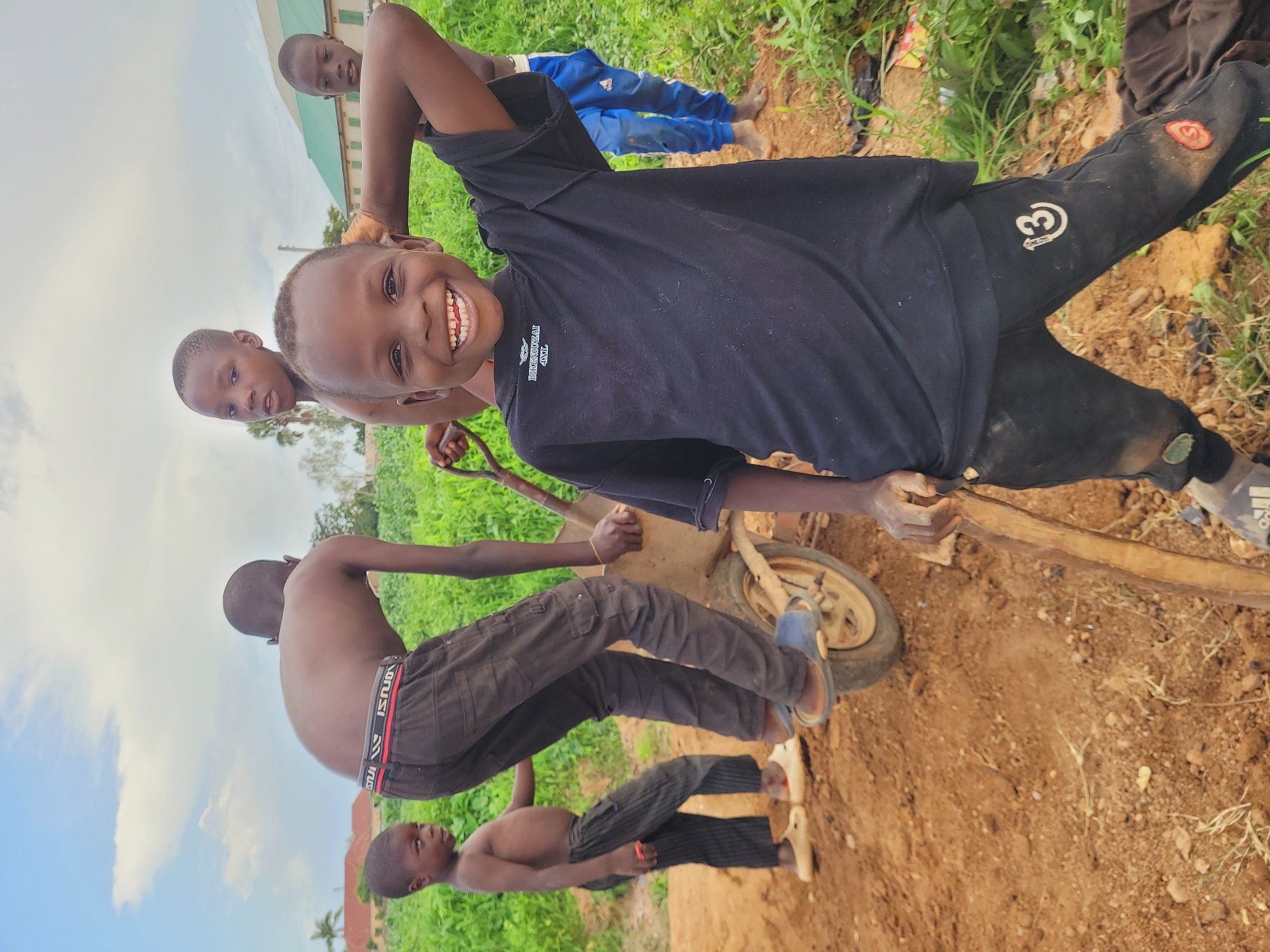 In Bukuru, Nigeria, under a partly cloudy sky, four young boys are engaged in activity around a wheelbarrow. The boy in the foreground, wearing a black t-shirt and dark pants with a "13" emblem, smiles broadly at the camera, his right arm raised. Behind him, another boy in dark pants and a red-striped waistband is bent over, interacting with the wheelbarrow. To the left, a shirtless boy in dark striped pants stands near the wheelbarrow. In the background, a fourth boy, wearing blue pants, stands partially obscured. The environment consists of loose dirt, green vegetation, and distant buildings.