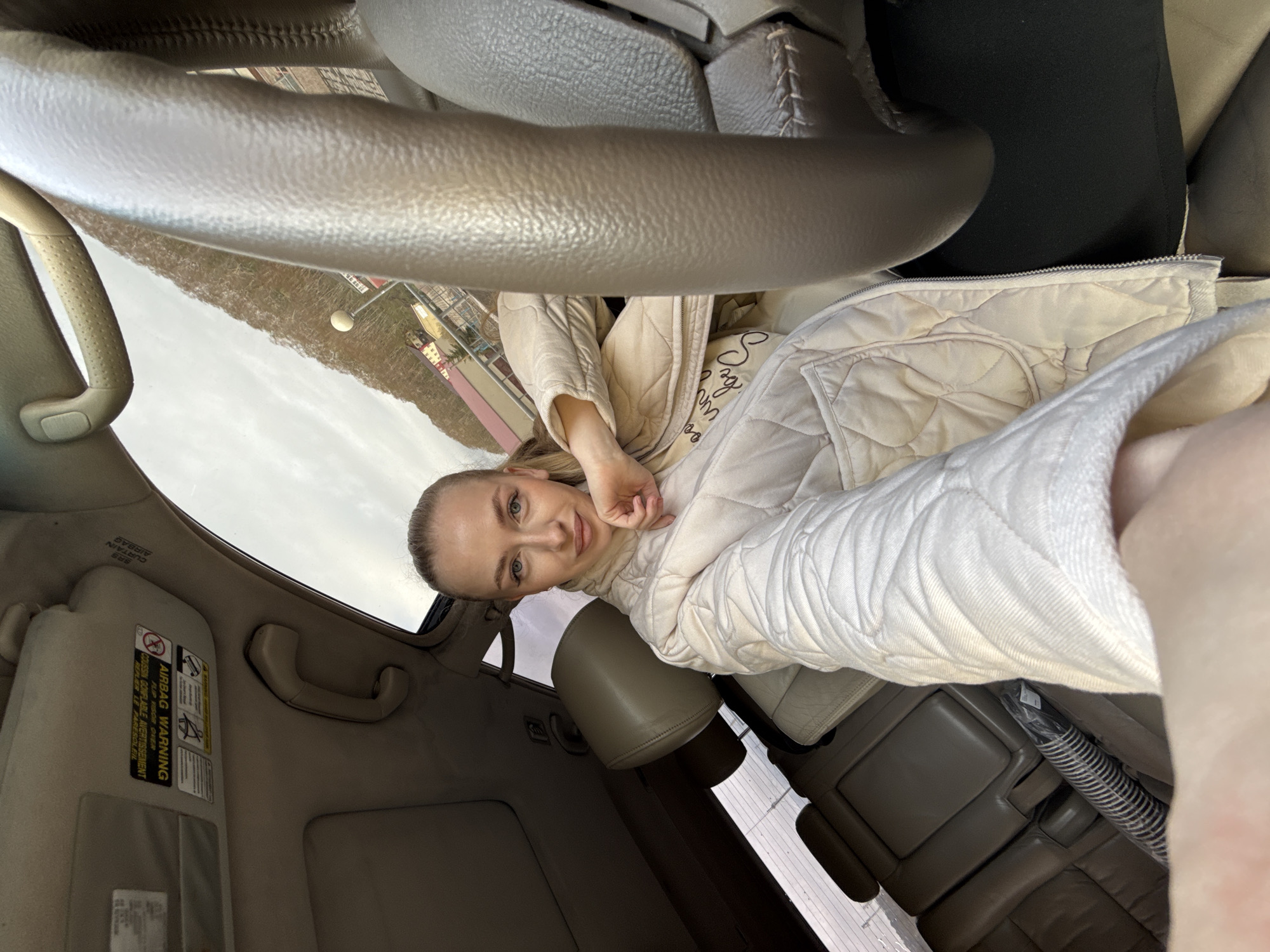 A blonde woman is taking a selfie in the driver's seat of a car. She is wearing a light-colored quilted jacket. The interior of the car is beige. The steering wheel is prominent in the foreground. Through the windshield, an urban landscape is visible, potentially suggesting Petropavlovsk-Kamchatsky, Russia. The car's sun visor with an airbag warning sticker is visible.