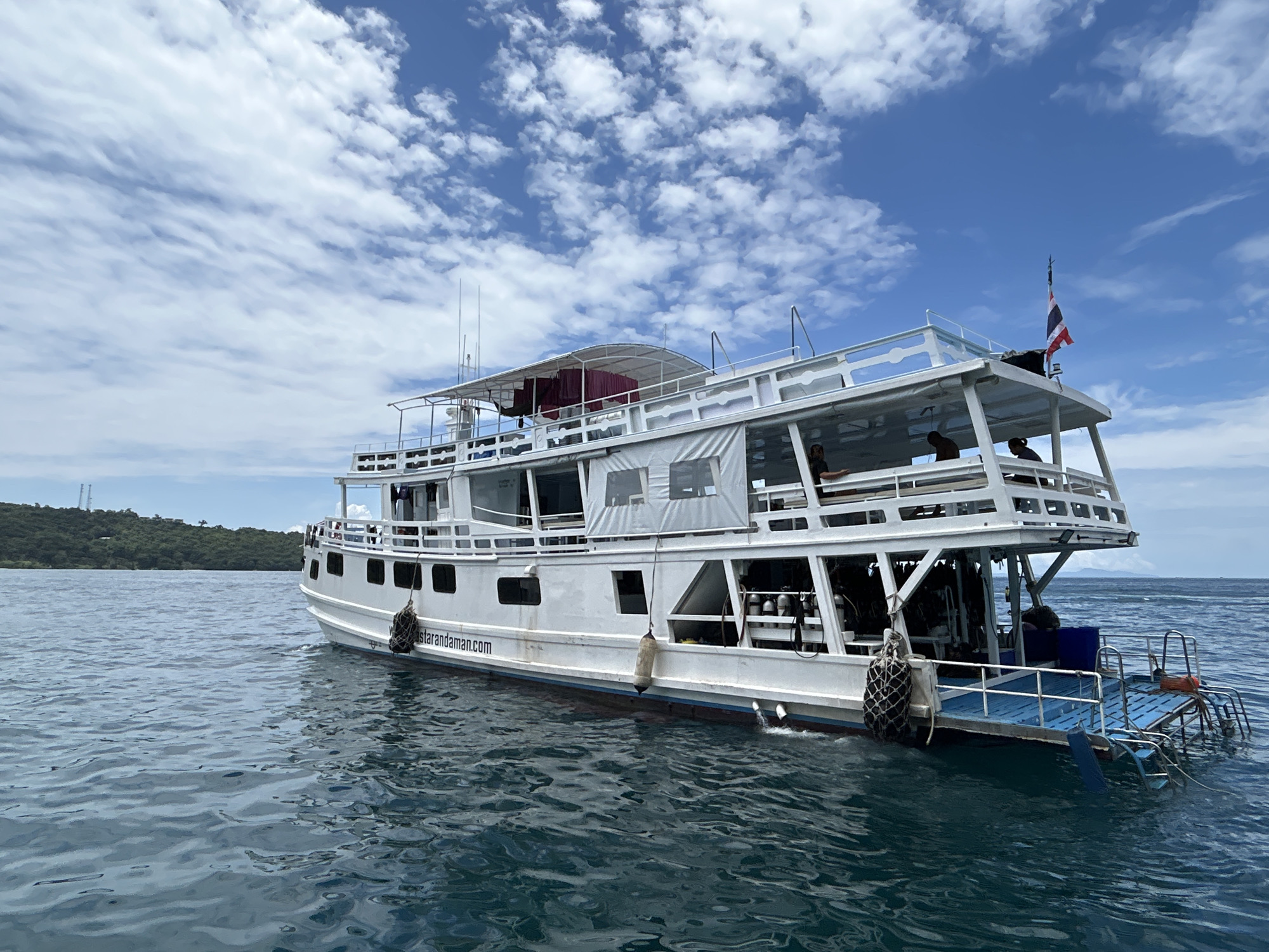 A white, multi-deck boat is moored in the water off the coast of Ao Nang, Thailand. The boat, which appears to be a dive boat, has several levels with open-air seating areas and enclosed cabins. There are scuba tanks visible on the lower deck, and a Thai flag is flying from the top deck. The boat is positioned at an angle to the camera, with the stern facing towards the right. The water is a deep blue-green, and the sky is a vibrant blue with scattered white clouds. In the background, a lush, green island can be seen rising from the sea. The overall impression is one of a peaceful, tropical setting.