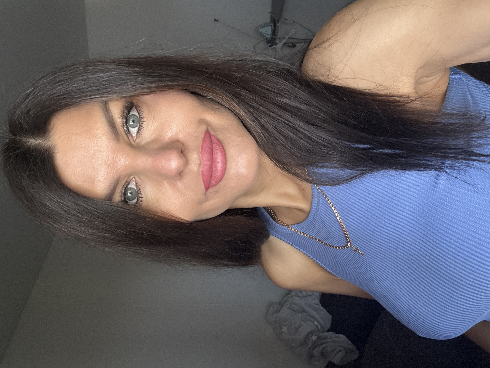 A medium close-up selfie of a woman with dark brown hair and striking blue-green eyes is shown. She is wearing a blue ribbed tank top and a rose gold chain necklace with a cross pendant. Her lips are painted a matte pink. The background is a dimly lit room with a gray wall on the left and what appears to be a dark couch with a grey blanket on the left side of the frame. A small electrical appliance or lamp is visible in the background on the right. The image is captured in Chaikovsky, Russia.