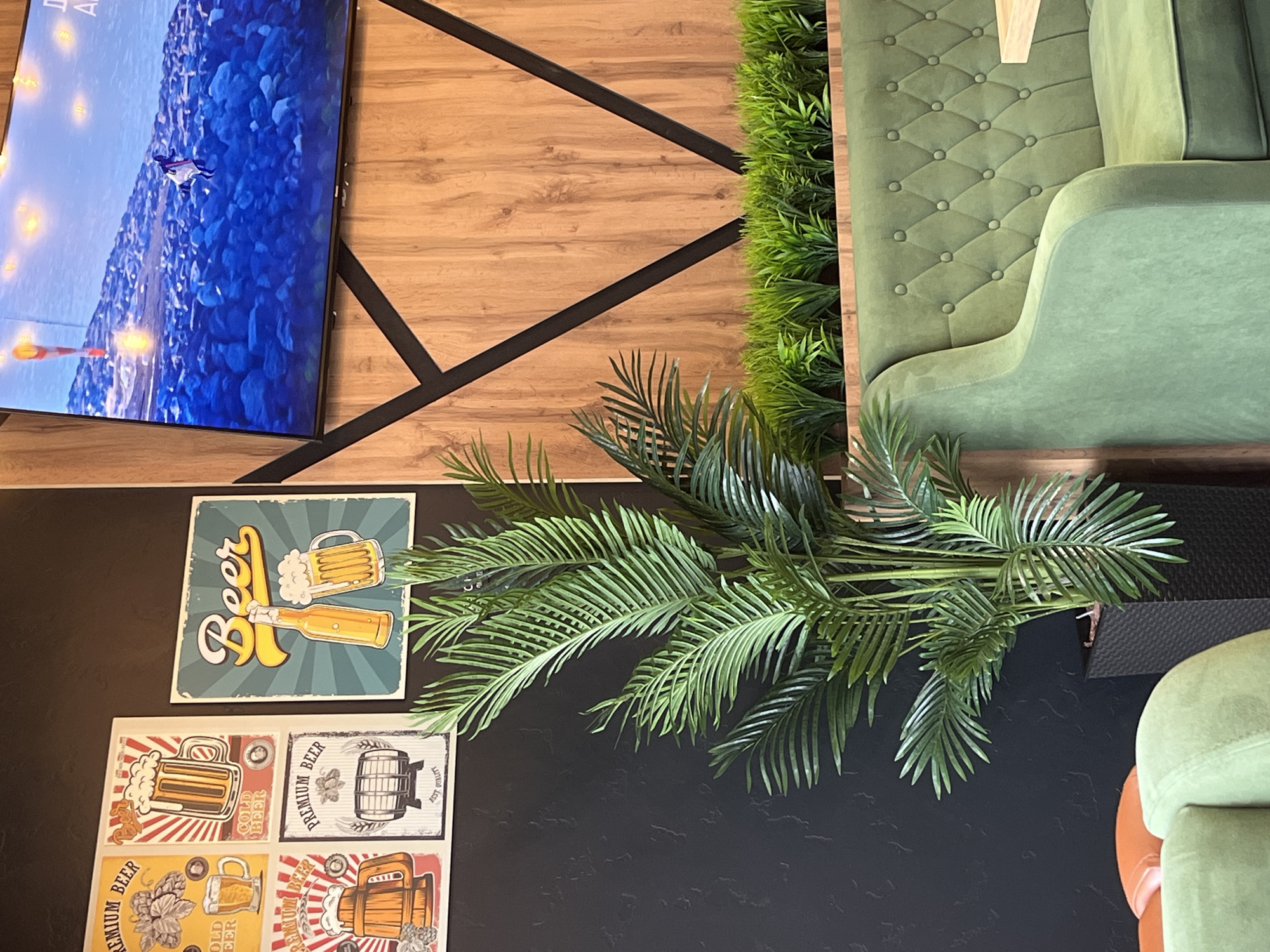 The image shows a section of a restaurant interior. On the left wall, there are four framed posters depicting beer and brewing imagery. To the right of these posters is a larger square poster with the word "Beer" written in a stylized font, accompanied by a drawing of a beer bottle and a beer mug. In the foreground, a large artificial palm plant is situated in a black rectangular planter. Behind the plant and the posters, a large television screen displays a scene of blue water and rocks, with a figure walking. The background wall features a wood-paneled design with black geometric accents. In the right portion of the image, a green upholstered booth seat with tufted detailing is visible, along with a portion of a light-colored table. A row of artificial greenery is planted behind the booth. The lighting is ambient, suggesting an indoor environment. The scene does not contain any identifiable individuals. The location is not specified.