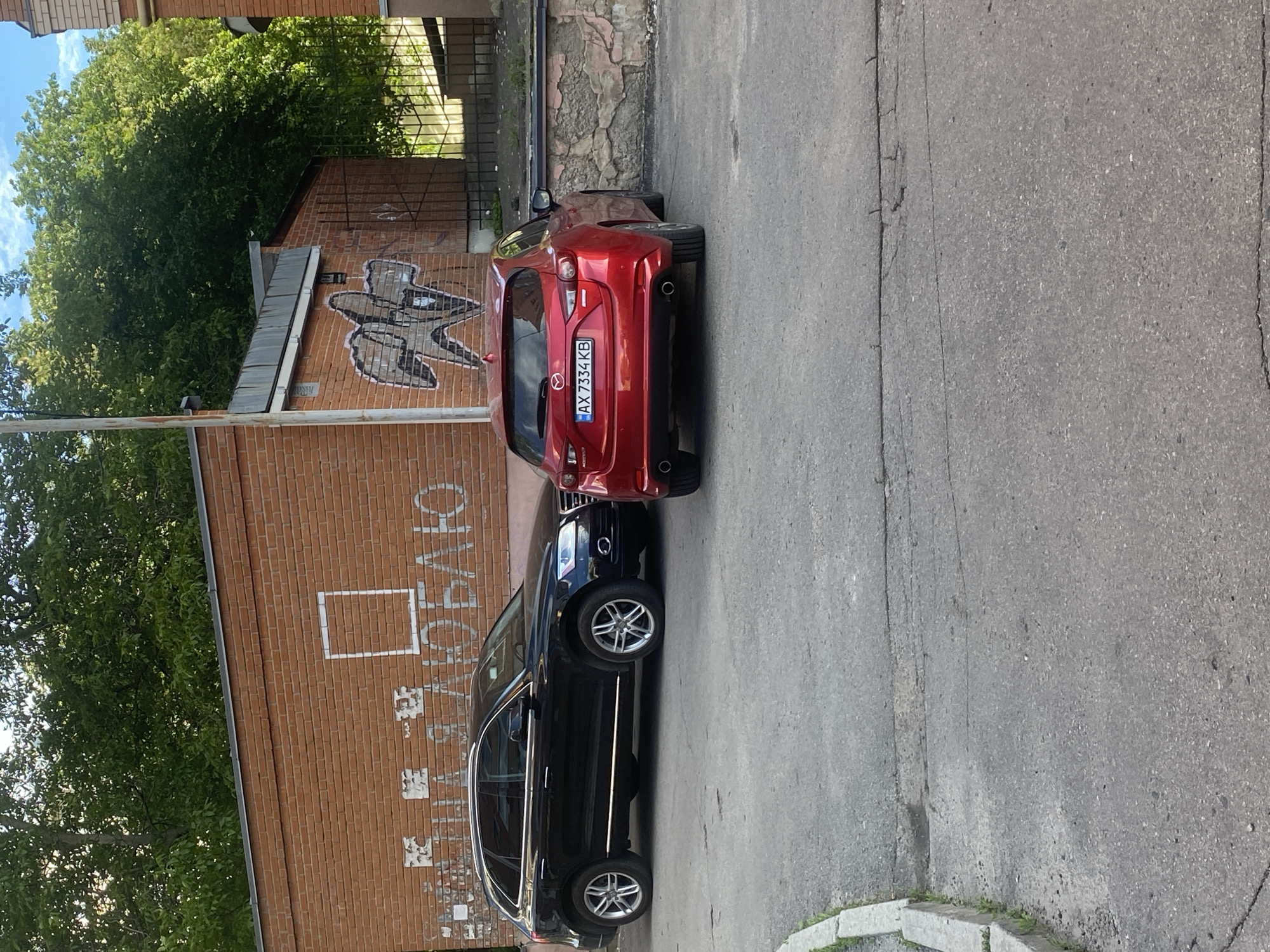 The image depicts a street scene in Poltava, Ukraine, featuring two parked cars. A red Mazda is parked directly behind a black Volvo wagon. The license plate of the Mazda is visible, displaying "AX 7334 KB." Both vehicles are parked along the side of a building constructed of red brick. Graffiti is visible on the brick wall, including a large stylized "M" or wing design, and the Cyrillic text "Полтаво" (Poltava). The surrounding environment includes trees, a section of sidewalk, and a portion of the street surface. The scene appears to be captured on a sunny day.