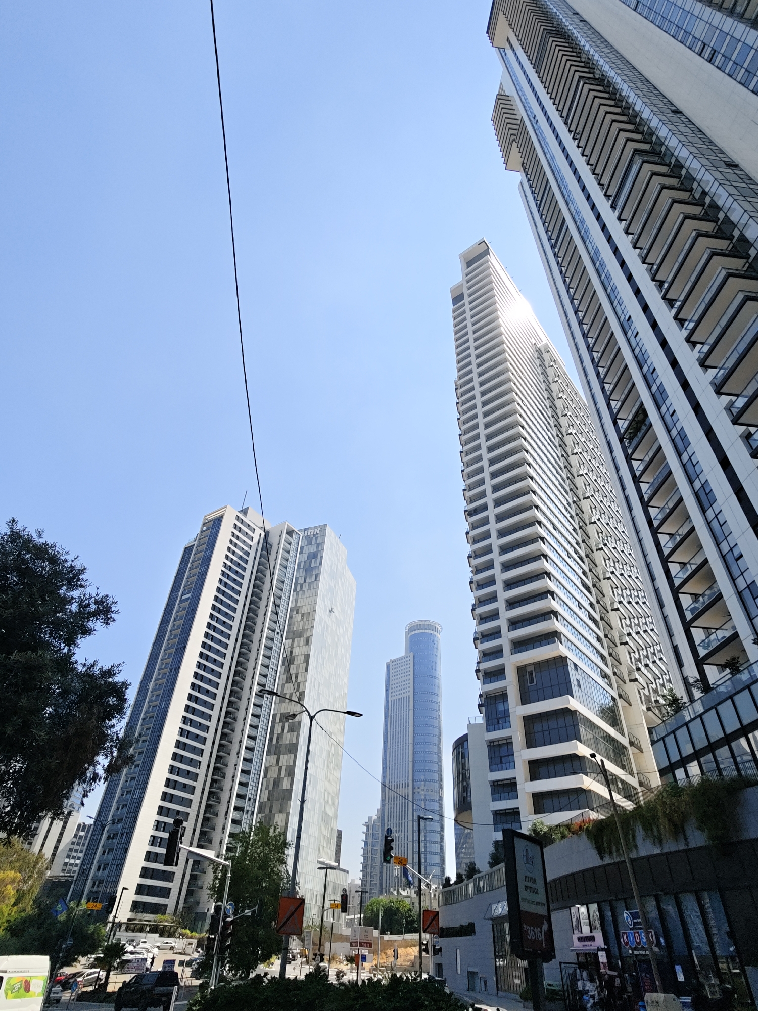 In Givatayim, Israel, a low-angle shot captures a clear blue sky dominating the upper portion of the frame. A thin, black wire runs diagonally from the top left to the mid-right. The lower half of the image is filled with several modern high-rise buildings. In the foreground to the left, the dark green foliage of a tree partially obscures the view. To the right of the tree, a cluster of tall, white buildings with numerous balconies and glass windows are visible, angled upwards towards the sky. Further to the right, another, even taller, white skyscraper with repeating balcony structures dominates the scene. In the mid-ground, a slimmer, blue-tinted cylindrical skyscraper stands out. The ground level shows a street with traffic lights, cars, and some pedestrians. The buildings are constructed with sleek, contemporary architecture.