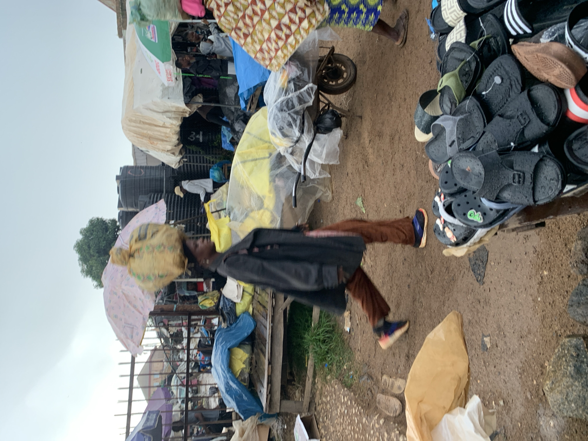 The image depicts a bustling outdoor market scene in Gura Top, Nigeria, under an overcast sky. In the foreground, a woman is walking from left to right, carrying a large yellow bundle on her head, partially covered by a pink umbrella. She is wearing a dark coat, brown trousers, and colorful sneakers. To her right, a wheelbarrow filled with items wrapped in plastic is visible. A pile of various sandals and flip-flops is also in the foreground, suggesting a stall or vendor. The background reveals more market stalls, tarpaulin-covered structures, large black water tanks, and other individuals engaged in market activities. The ground is muddy and appears to be unpaved. The overall atmosphere suggests a busy commercial area, likely operating in challenging weather conditions.