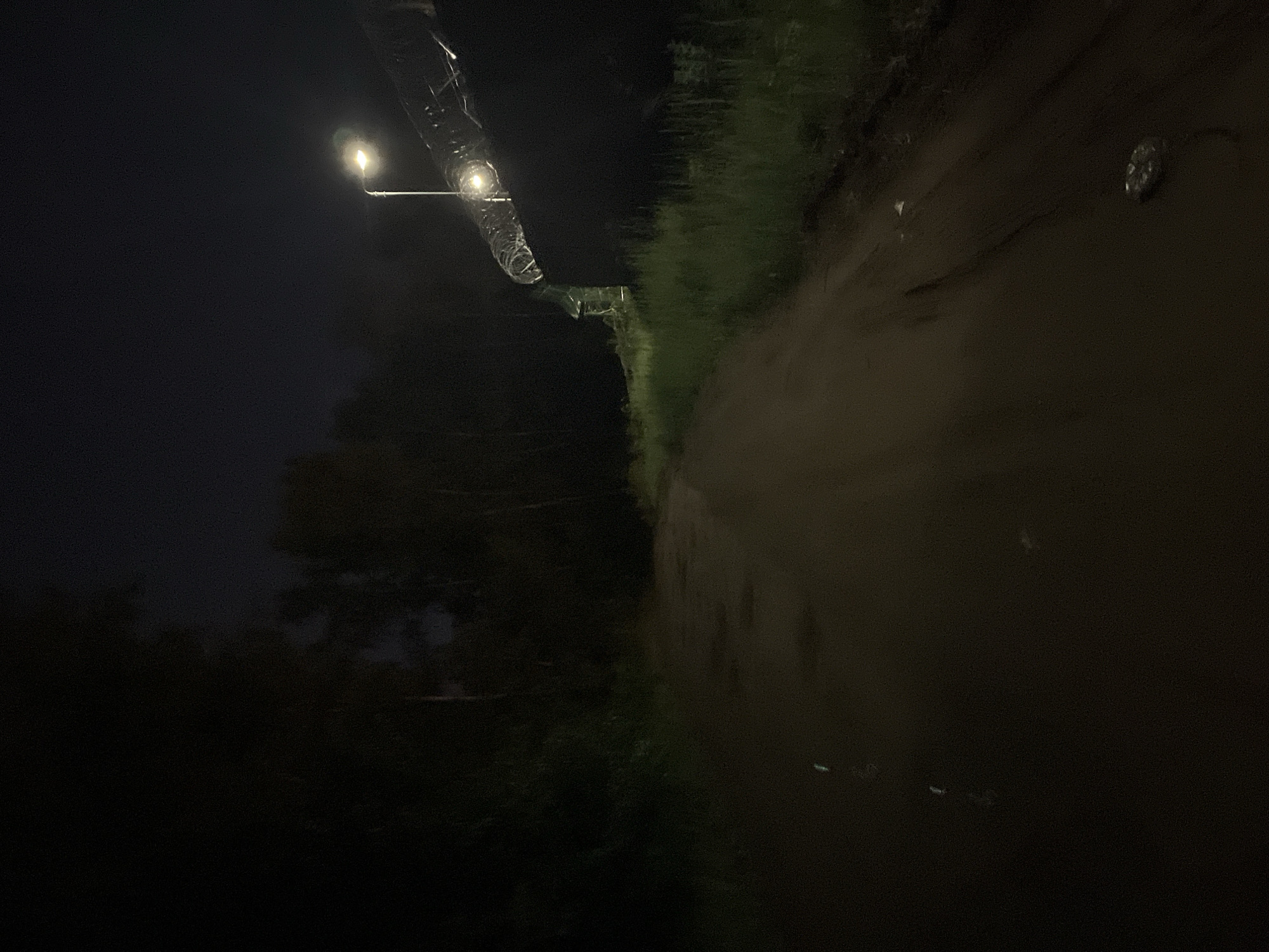The image depicts a nighttime scene along an unpaved road in the city of Chaikovsky, Russia. The road, composed of dark earth, occupies the lower and central portion of the frame, showing textural variations and subtle reflections or light spots, particularly towards the bottom right and center-left. To the left, dense, dark foliage and trees are visible, silhouetted against the dim sky and extending into the background.
On the right side of the road, a dark, solid structure or wall runs parallel to the path. This structure is topped with a section of coiled razor wire (barbed wire), brightly illuminated by an overhead street light. Green grass grows between the unpaved road and the base of this wall.
A single street light, mounted on a pole, is positioned above the right side of the path, casting a localized, yellowish-white light onto the razor wire, the upper part of the wall, and a segment of the grass and road. The light source itself emits a soft glow. The sky above is dark, with faint indications of clouds or haze. No human figures, animals, or explicit actions are discernible within the scene. The environment suggests an outer or industrial edge of a developed area due to the combination of unpaved road, dense vegetation, and security fencing.