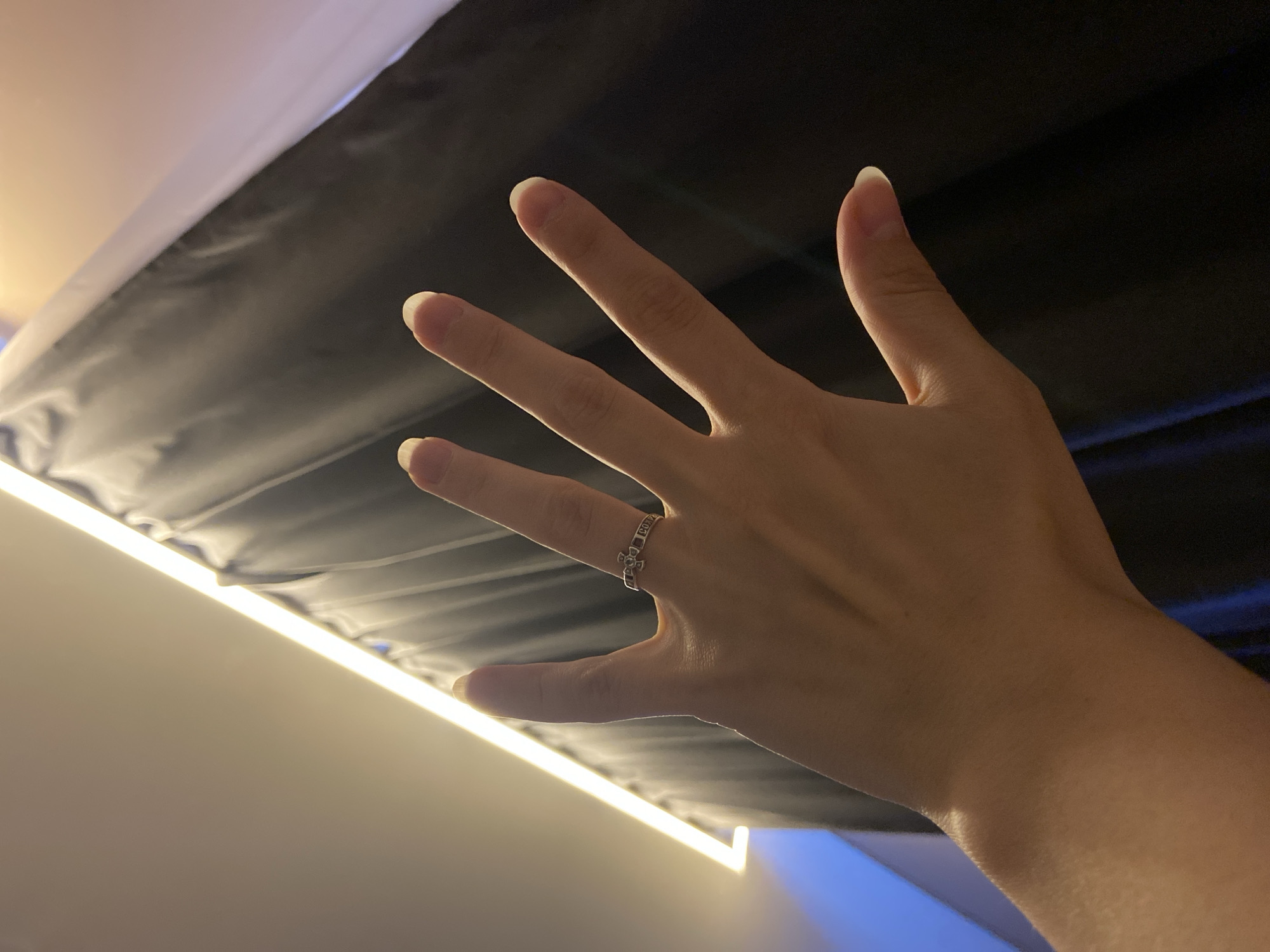 The image, taken in Peresvet, Russia, features a person's hand extended upwards with the palm facing the camera. A silver-colored ring, adorned with an intricate pattern of connected small crosses or squares, is visible on the ring finger.
A prominent, horizontal linear light source, likely an LED strip, runs across the midground, illuminating the hand and parts of the background. Above this light, a dark, textured surface, possibly fabric or the underside of a furniture piece, fills the upper section of the frame, showing subtle folds. Below the linear light and behind the hand, a lighter, off-white surface is present, and a soft blue glow emanates from the bottom right corner, suggesting another ambient light source or a screen.
The photograph is captured from a low-angle perspective, highlighting the upward pose of the hand within an indoor setting. The artificial lighting suggests the time is either night or a controlled lighting environment. No specific activity beyond the hand's pose is discernible, and no explicit text is clearly visible on the ring or elsewhere in the scene.
