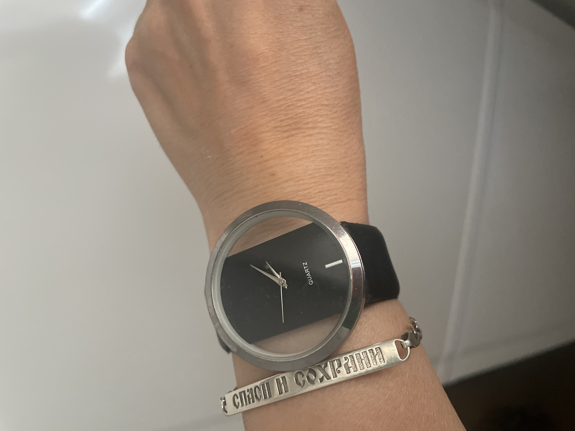 A close-up shot focuses on a person's left wrist, adorned with a modern watch and a silver bracelet. The watch features a large, transparent face with a minimalist black dial and a black strap. The hands of the watch are silver, pointing to approximately 10:10. The word "QUARTZ" is visible on the dial. Below the watch, a silver bracelet is worn. The bracelet has a rectangular charm with engraved text in Cyrillic letters that appears to read "СПАСИ И СОХРАНИ" (Save and Protect). The background is a plain, light-colored surface, possibly a wall or countertop, with some indistinct shadows. The lighting suggests it's daytime. The image is shot in Almaty, Kazakhstan. No specific emotions are conveyed, and the activity is simply wearing accessories.