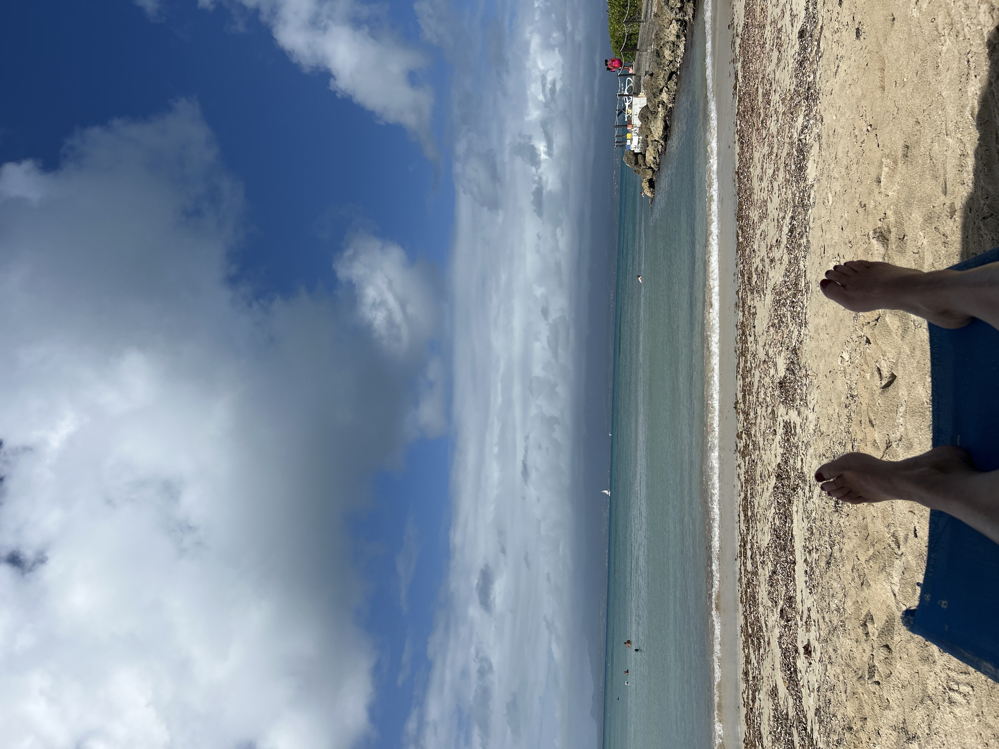 The image depicts a beach scene in Le Gosier, Guadeloupe, France. The view is from the perspective of someone lying on a blue beach chair, with their feet visible in the lower right corner of the frame. The beach is sandy, with visible seaweed or debris scattered across the surface. The water is calm and turquoise in color, with small waves breaking along the shore. Several people are visible in the water, suggesting swimming or other water activities. In the background, there is a rocky outcrop with some structures on top, possibly a lifeguard station or a small building. The sky is partly cloudy, with patches of blue visible between the clouds. The image is oriented vertically.