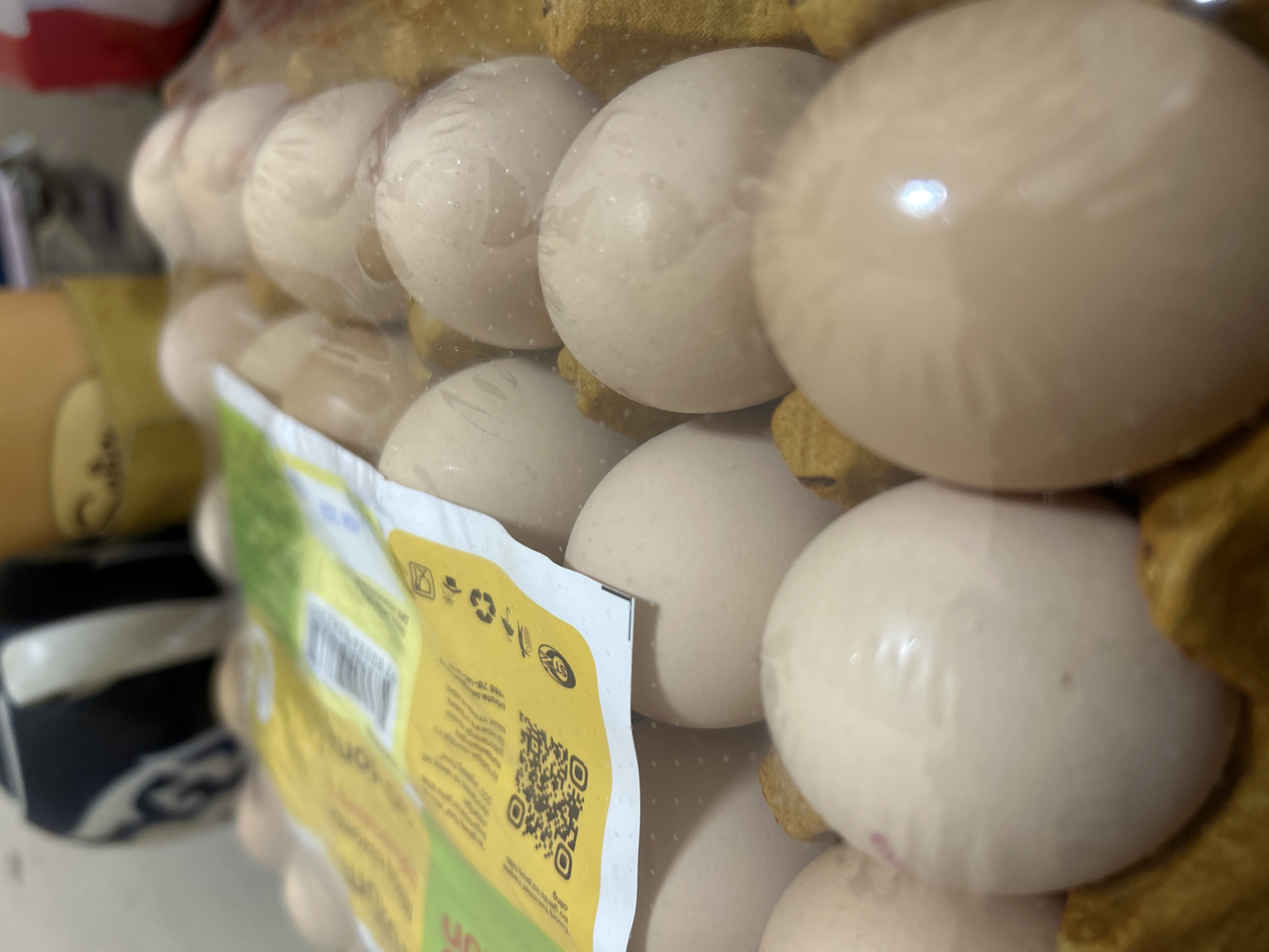 A close-up, slightly blurry, indoor shot shows a carton of pale, speckled eggs. The eggs are wrapped in clear plastic, and some have a pink stamp or mark on them. A yellow and green paper label with a QR code and some text is folded over the top of the egg carton. The background is out of focus, but appears to be a domestic setting with some objects on shelves. The image is not specific to Ahmad Yasawi, Uzbekistan.