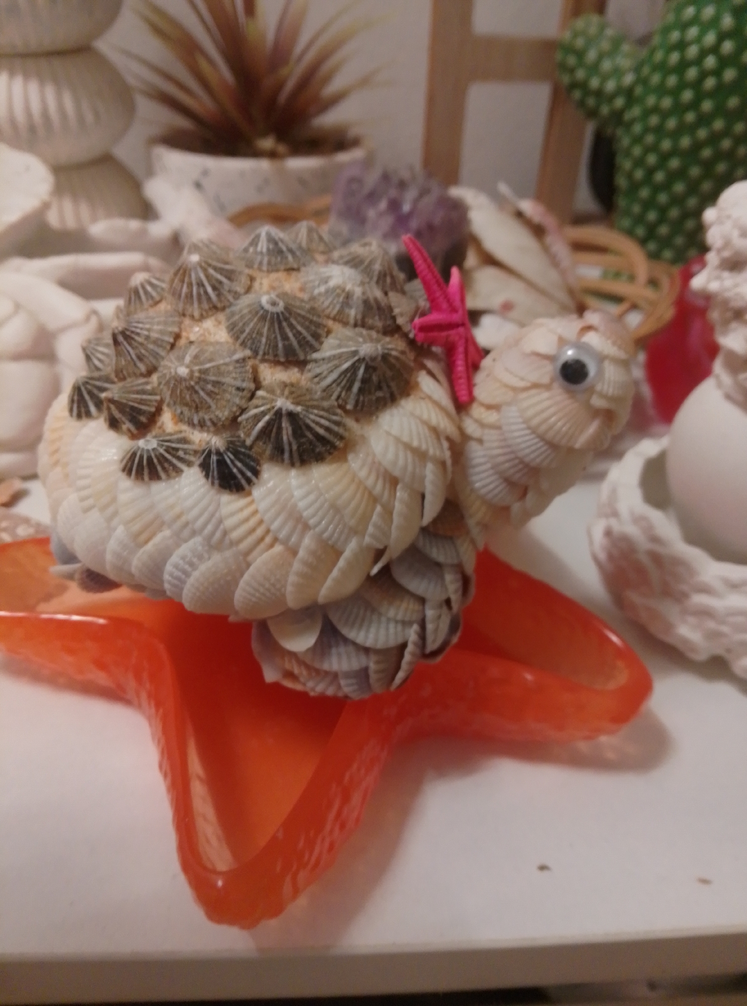 The image shows a handmade turtle figurine crafted from seashells. The turtle is placed on a glossy orange starfish-shaped dish. The figurine has a detailed shell made of various small shells and has googly eyes. A small pink starfish decoration is attached to its back. In the background, out of focus, are other decorative items, including a small potted plant, what appears to be a cactus figurine, and other seashell decorations. The scene is set on a white surface, possibly a table or shelf. The image does not provide any information about the city of Wedel, Germany.