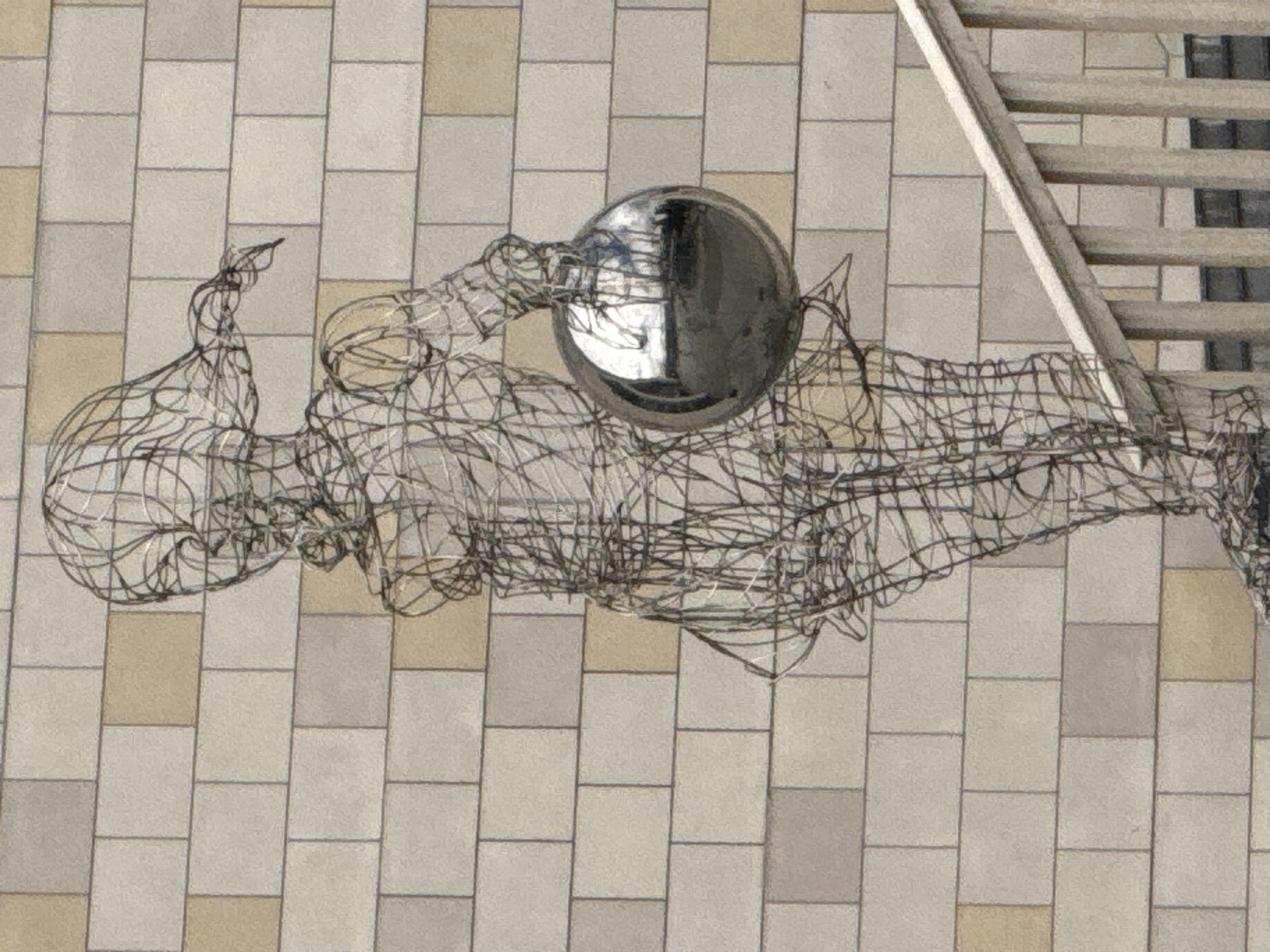 The image shows a wireframe sculpture of a young girl holding a large, reflective sphere. The sculpture is positioned in front of a beige tiled wall. A wooden railing is visible in the foreground, extending from the bottom right of the frame upwards. The girl's body is constructed from thin, interwoven wires, creating an airy, translucent effect. Her hair is depicted as flowing strands of wire, and she wears a simple dress. The sphere she holds is highly polished, reflecting the surrounding environment, which appears to include trees and possibly a building. The lighting suggests it is daytime, with soft shadows cast by the sculpture and railing. The overall scene is of an outdoor public space, likely a park or plaza, given the presence of art and architectural elements. There is no indication of the specific location within Moscow, Russia.