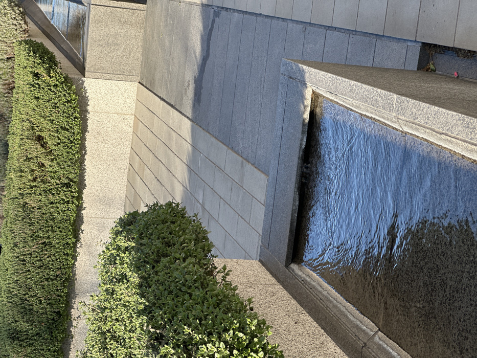The image displays a modern, tiered urban landscape feature, specifically located in Canada Square Park, Canary Wharf, London, United Kingdom. In the foreground, a rectangular granite water channel, dark gray on its interior, contains flowing, rippling blue water. Water cascades into this channel from a hidden source on the left. Parallel to the water channel, a granite planter on the left contains a dense, manicured green hedge. The midground features a paved walkway composed of two distinct sections of rectangular paving slabs: one of lighter gray and an adjacent section of darker gray, both laid parallel to the water feature. In the background, a higher granite planter wall supports a second level of dense green hedging. The scene is illuminated by natural sunlight, casting defined shadows on the paving and water, indicating a clear day. No individuals or animals are present.