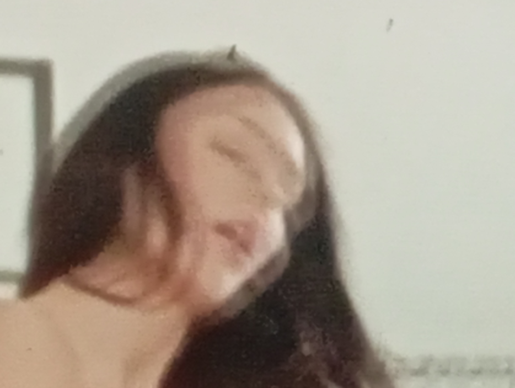 The image is a close-up, out-of-focus photograph of a person's face and upper body. The subject has long, dark hair that frames their face. Their eyes are closed, and their head is tilted downwards. The background is a plain, light color, possibly a wall. There are no identifiable landmarks or contextual elements to indicate a specific location within Sofiivska Borshchahivka, Ukraine. The image quality is poor, with low resolution and blurred details, making it difficult to ascertain further specifics about the subject or their surroundings.