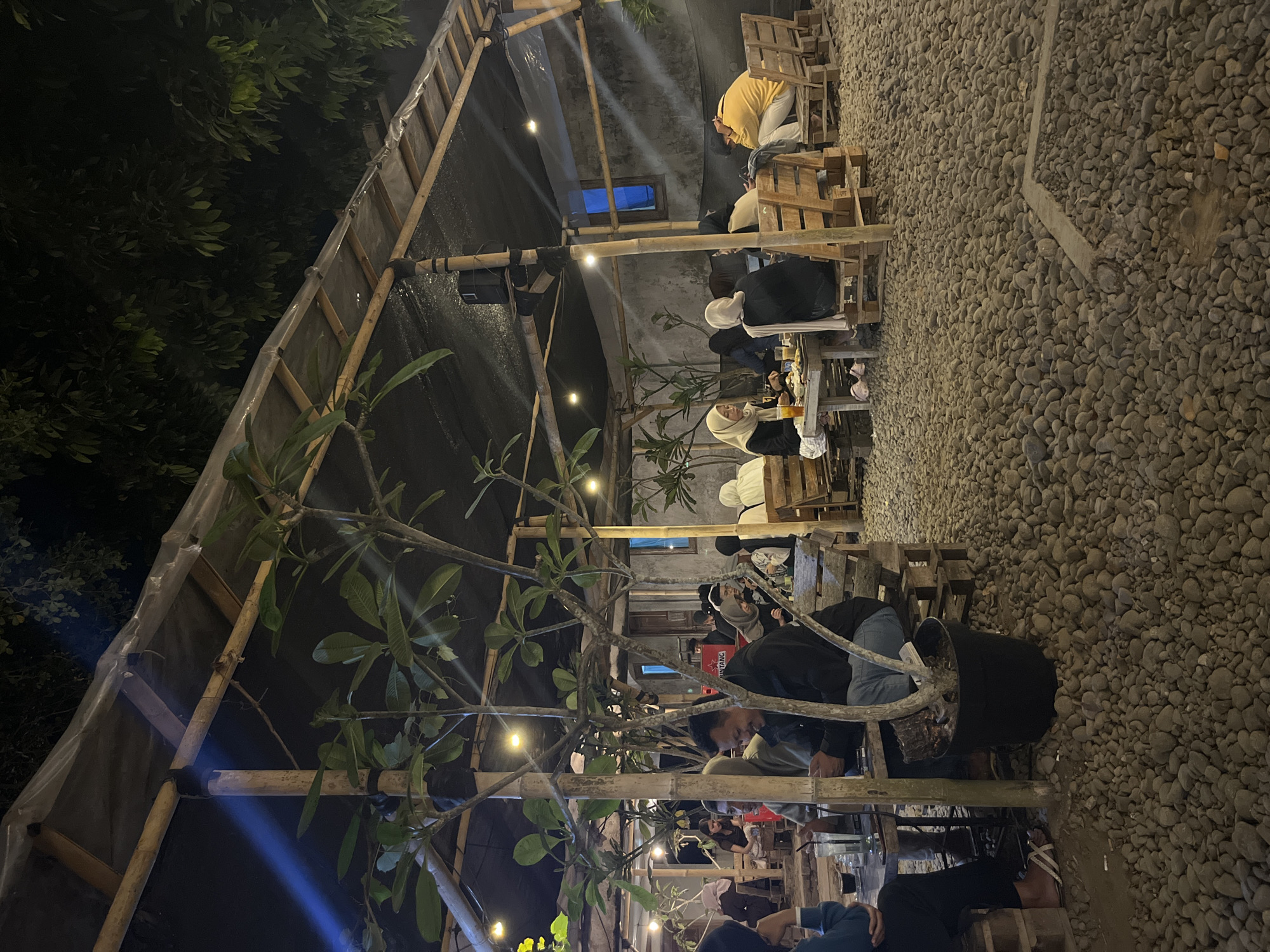 The image depicts an outdoor seating area at night, likely a cafe or restaurant, in Matesih, Indonesia. The area is covered by a makeshift roof constructed from bamboo poles and dark tarpaulin. String lights are strung across the canopy, providing illumination. The ground is covered with small, grey pebbles.
Several groups of people are seated at tables. Most are dressed in casual attire, with some women wearing headscarves. They appear to be socializing and eating or drinking. The furniture consists of tables made from what looks like recycled wooden pallets and benches also constructed from pallets. A potted plant is situated in the foreground, next to a bamboo support pole. The background shows a concrete wall with some windows. The overall ambiance is relaxed and informal, suggesting a popular local gathering spot.