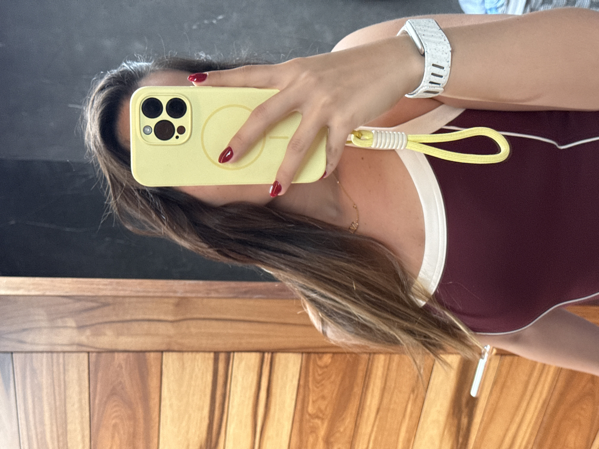 A person is holding a yellow smartphone with a camera lens area containing four lenses and a wrist strap in front of their face, obscuring most of it. The person has long, light brown hair and is wearing a burgundy top with white trim. A white smartwatch is visible on their wrist. The background includes a wooden surface, possibly a wall or furniture, and a blurry dark gray or black area, suggesting a mirror or a darker space. The location of San Bernardino Tlaxcalancingo, Mexico cannot be confirmed from the visual information provided.