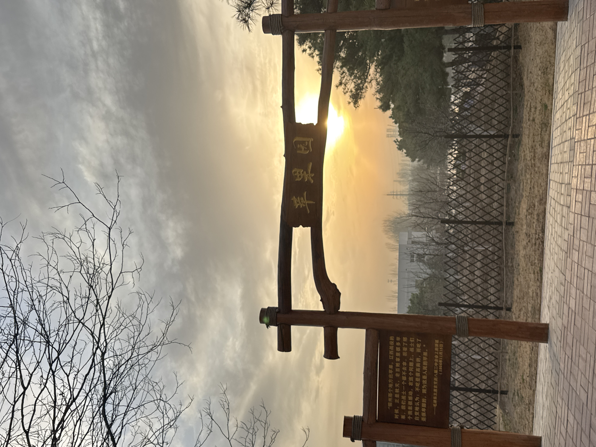 The image captures a wooden archway at the entrance to Apple Garden in Zhongtun, China, against a backdrop of a bright, hazy sunset. The wooden structure consists of two upright posts supporting a horizontal beam. A signboard bearing golden Chinese characters hangs from the beam. In the background, trees and a fence are visible, along with the silhouette of buildings in the distance. A leafless tree stands to the left of the archway. A paved path leads into the park. To the right of the archway is another sign with printed text in Chinese.
