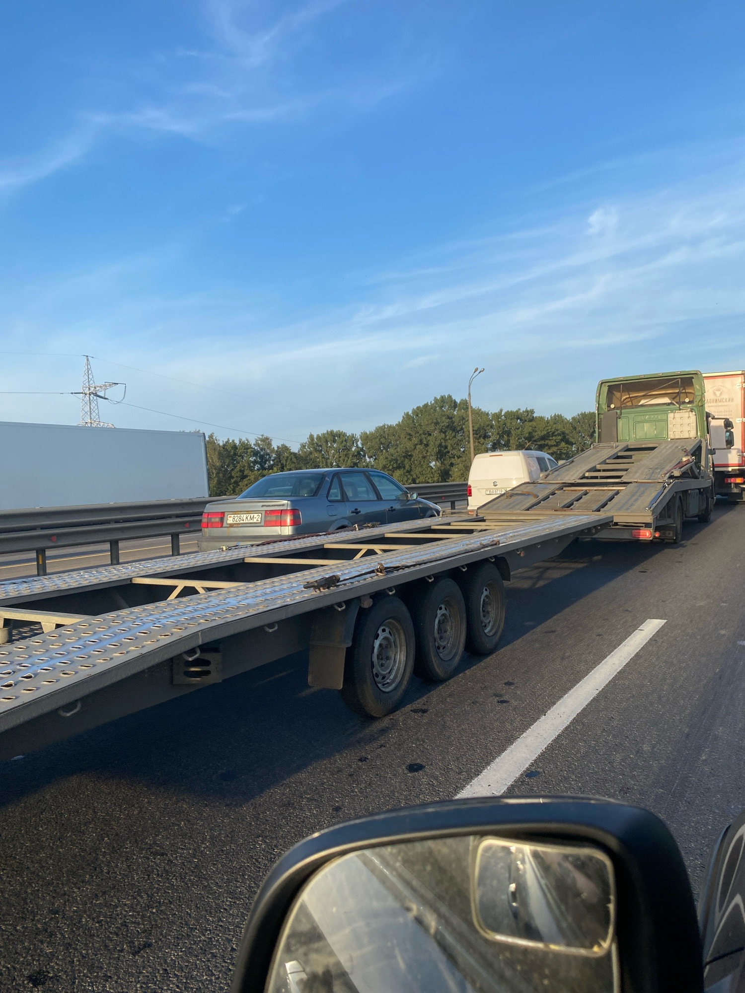 A vehicle is being transported on a flatbed truck on a highway. The truck is green and has a long, empty trailer. In the background, there are trees, a power line tower, and a blue sky with white clouds. A silver car is on the highway to the left of the truck. The image appears to be taken from inside another vehicle, as a side mirror is visible in the foreground. The license plate of the silver car is visible and reads "98284 KM-2". The license plate of a white van behind the truck reads "9719". The scene is set on a highway in Minsk, Belarus.