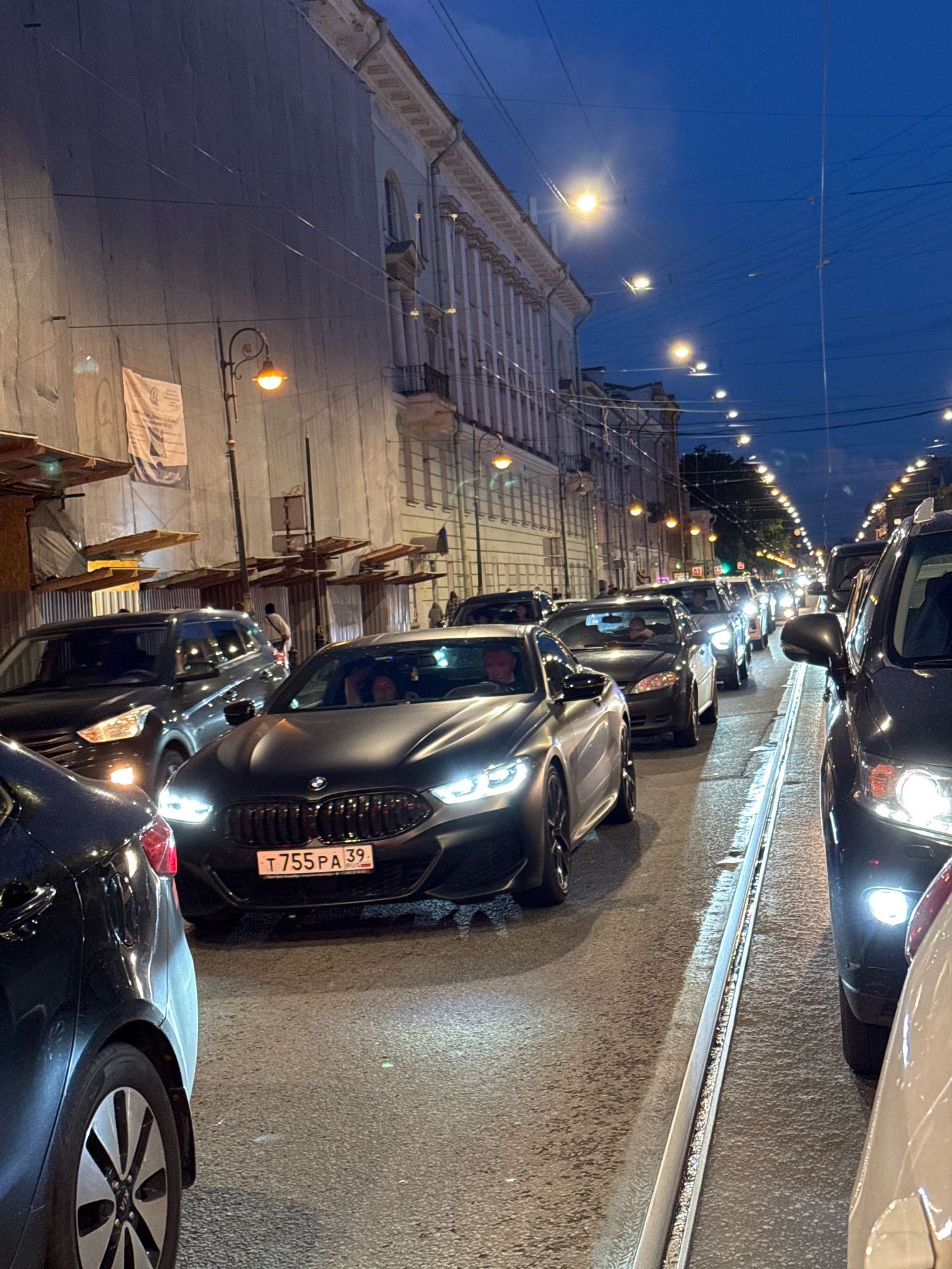 The image depicts a nighttime traffic jam on a city street, likely in Saint Petersburg, Russia, given the architectural style and the license plate format. The foreground features the rear of a dark-colored vehicle, partially obscuring the view. Immediately behind it is a black BMW with its headlights illuminated, displaying the license plate "T 755 PA 39". Inside the BMW, several individuals are visible.
The street is lined with various cars, all stationary in traffic. The buildings on the left are tall and appear to be old, with classical architectural elements. They are partially covered by scaffolding or tarps, suggesting ongoing construction or maintenance. Streetlights are visible along the street, casting an orange glow. The sky is dark blue, indicating dusk or nighttime. Tram tracks run along the center of the road, embedded in the asphalt. The overall atmosphere is one of congestion and urban evening.