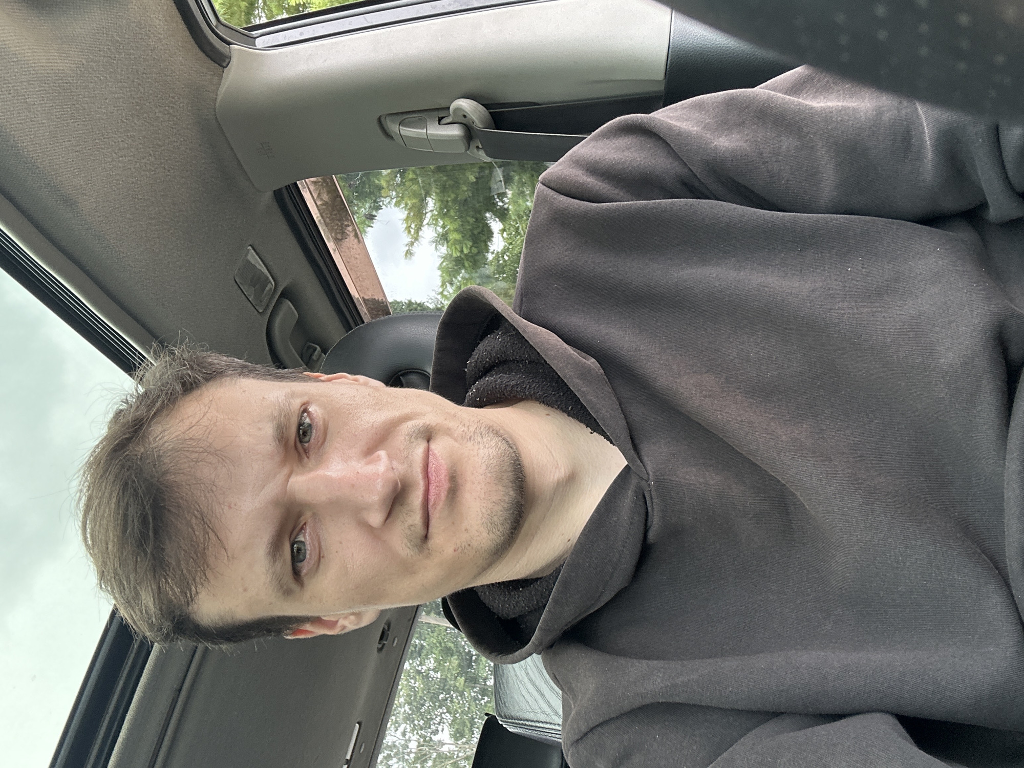 A close-up, indoor, selfie-style photograph features a man with light brown hair and green eyes wearing a dark brown hooded sweatshirt. He is seated inside a vehicle, with a sunroof visible above him. The background shows greenery through the car window.