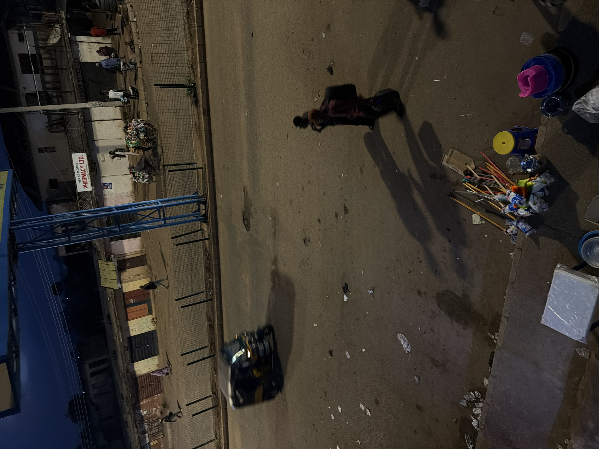 This image depicts a street scene in Bukuru, Nigeria, at dusk or night. In the foreground, various items are scattered on the ground, including what appear to be brooms, colorful sticks, plastic containers, and some debris. A light blue and yellow stool is visible, along with a stack of colorful plastic bowls and a smaller grey ashtray. A person is walking away from the camera in the middle ground, casting a long shadow, carrying what appears to be luggage on their back. Further in the distance, a three-wheeled auto-rickshaw is captured in motion, its headlights on, also casting a shadow.
Across the street, a row of shops or market stalls is visible. One stall has a sign that reads "NERAT MODEL PHARMACY LTD." Other stalls have closed doors and windows. Several people are seen near the shops, some standing and some walking. One individual is pushing a cart laden with goods. The street itself is paved and appears somewhat littered. Overhead, power lines are strung across the scene, and a large structure, possibly a billboard or part of a building, is visible at the top of the frame. The overall atmosphere suggests a busy urban environment transitioning from day to night.