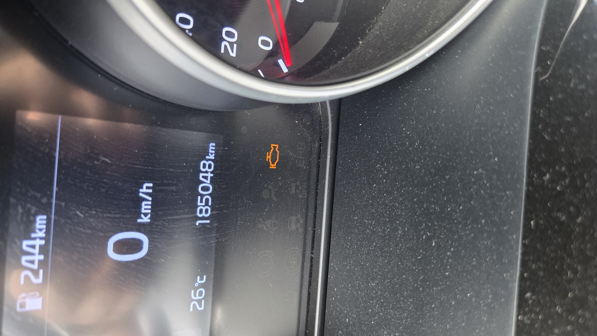 The image shows a close-up view of a car's dashboard, specifically the digital display and part of the speedometer. The display shows several pieces of information:
* **Fuel Gauge:** Indicates 244 km remaining.
* **Speed:** Reads 0 km/h.
* **Temperature:** Shows 26°C.
* **Odometer:** Reads 185,048 km.
* **Check Engine Light:** An orange icon of an engine is illuminated, indicating a potential problem with the vehicle.
* **Other Icons:** Several other warning or indicator lights are partially visible, including what appears to be ABS, traction control off, and seatbelt indicators.
The dashboard is dusty. The speedometer on the right is partially visible, with the needle pointing to zero.
The image does not provide any information about the city of Burgas, Bulgaria, or any specific environmental details that would place it in that location.