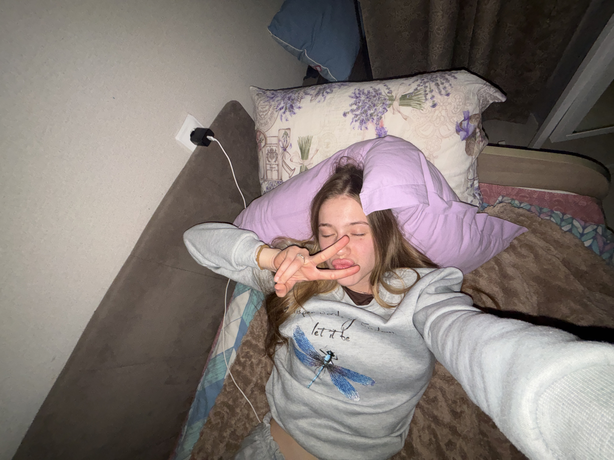 The image shows a young woman lying on a bed, possibly in Ladozhskoe Ozero, Russia. She is wearing a light gray sweatshirt with a blue dragonfly graphic and text. Her eyes are closed, and her tongue is sticking out slightly. She is holding up two fingers in a peace sign gesture near her face. The bed is covered with a brown textured blanket and a patterned sheet. To her right, there is a light purple pillow and another pillow with a lavender floral pattern. Behind the pillows, a blue pillow is visible. The background shows a sloped ceiling and a wall with an electrical outlet and a plugged-in device. The scene appears to be indoors, in a domestic setting.