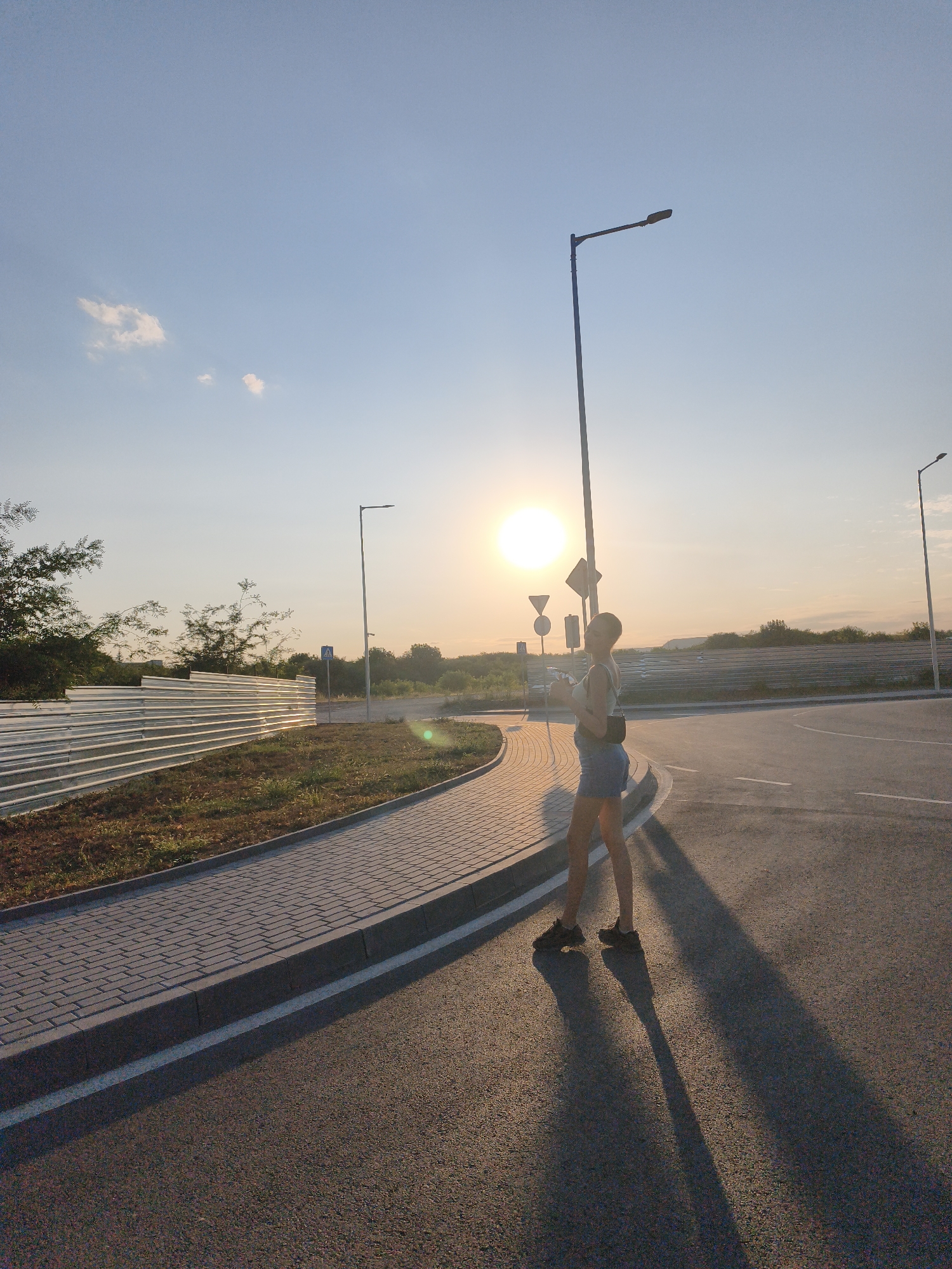 The image captures a young woman standing on an asphalt road near a paved walkway, bathed in the warm glow of a setting or rising sun. She is wearing a light-colored top and denim shorts, with a dark shoulder bag. Her shadow stretches long across the road. In the background, a tall street lamp stands prominently, and a metal fence separates the road from a grassy area with scattered trees. Traffic signs are visible on poles. The sky is clear with a few wispy clouds, and the sun is positioned low on the horizon, casting a bright light and a warm hue over the scene. The location is the city of Lovech, Bulgaria.