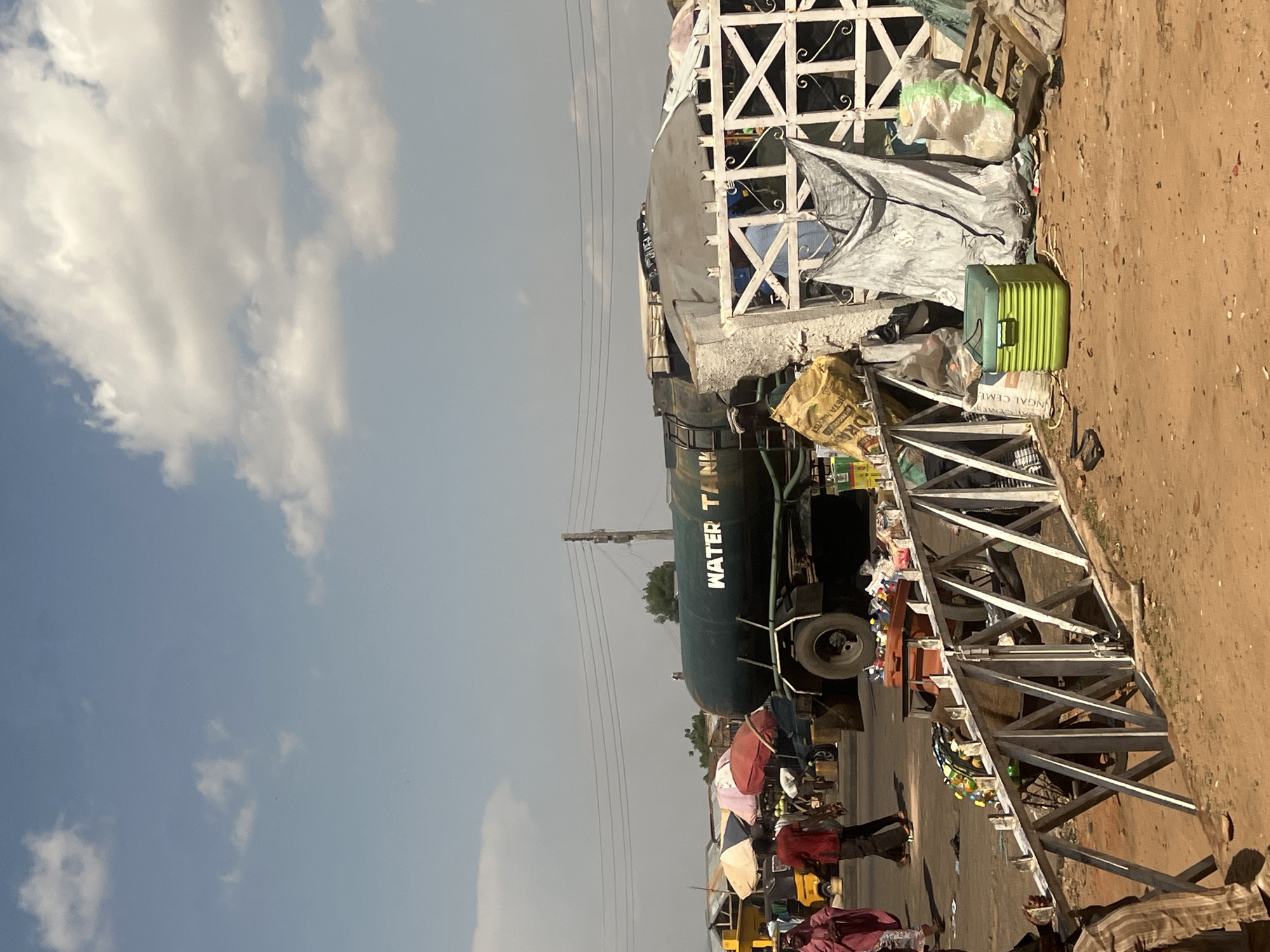 This is a daytime street scene in Gombe, Nigeria. The sky is light blue with scattered white cumulus clouds. The ground primarily consists of reddish-brown dirt and sand, with some asphalt visible on a road surface in the middle ground.
In the foreground, a low, white, metallic barrier with crisscross and rectangular patterns runs horizontally. To the right of this barrier, on the ground, are various items including a bright green cooler box, multiple plastic bags, a large grey tarp hanging from a structure, a light brown sack with "BETA HARVEST" visible, and several wooden pallets. A person in striped trousers is partially visible sitting on the far left, obscured by the barrier.
In the mid-ground, a dark green water tanker truck with "WATER TANK" painted in white capital letters on its side is a prominent feature. The truck is positioned on what appears to be a paved road. To the left of the tanker, several individuals are walking; one person wears a pink garment, and another wears a red shirt and dark trousers. Behind these individuals, a yellow auto-rickshaw is partially visible, along with multiple market stalls or temporary shelters topped with large umbrellas (white, red, and other colors). A dark vehicle is partially visible behind the tanker truck.
In the background, overhead power lines stretch across the frame, connected to a utility pole on the right. Distant buildings and structures are faintly visible under a somewhat hazy sky.