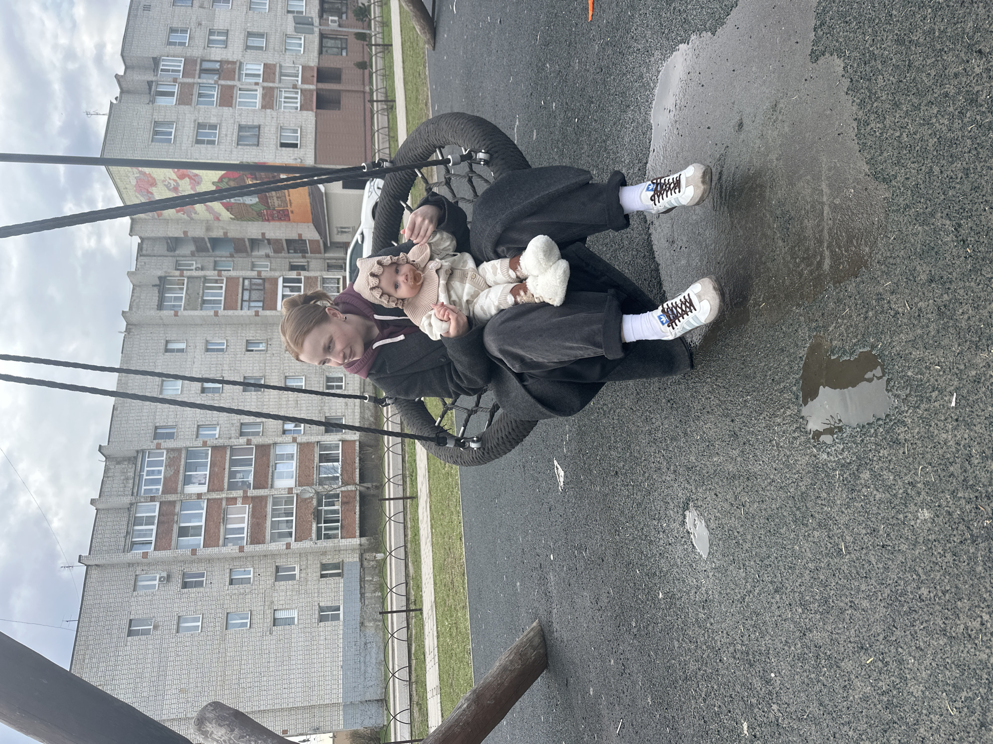The image shows a woman and a baby on a tire swing in a playground in what appears to be Pokrov, Russia, judging by the architecture. The woman is wearing dark trousers and light sneakers with white socks. The baby is bundled in a light-colored outfit with a bonnet and has a pacifier in its mouth. They are sitting in the tire swing, which is suspended by thick black ropes.
The background features a large, multi-story residential building with a grid of windows and a mural on its side. The playground surface is asphalt, with a puddle of water visible. The overall lighting suggests an overcast day.