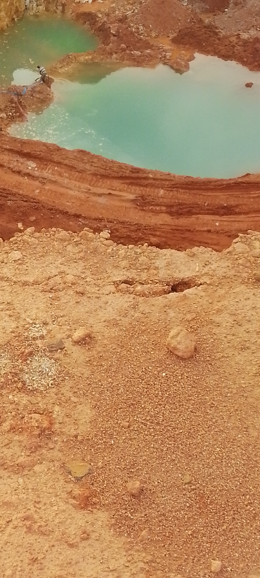 An aerial perspective of a partially excavated quarry pit filled with turquoise water. The pit's edges are defined by exposed layers of reddish-brown earth and rock. A single individual, wearing a striped shirt and dark pants, stands on a mound of earth near the edge of the water. The foreground consists of a dry, sandy area with scattered rocks and pebbles. The overall scene suggests an active or recently active mining or construction site. The location is identified as Unknown, Unknown.