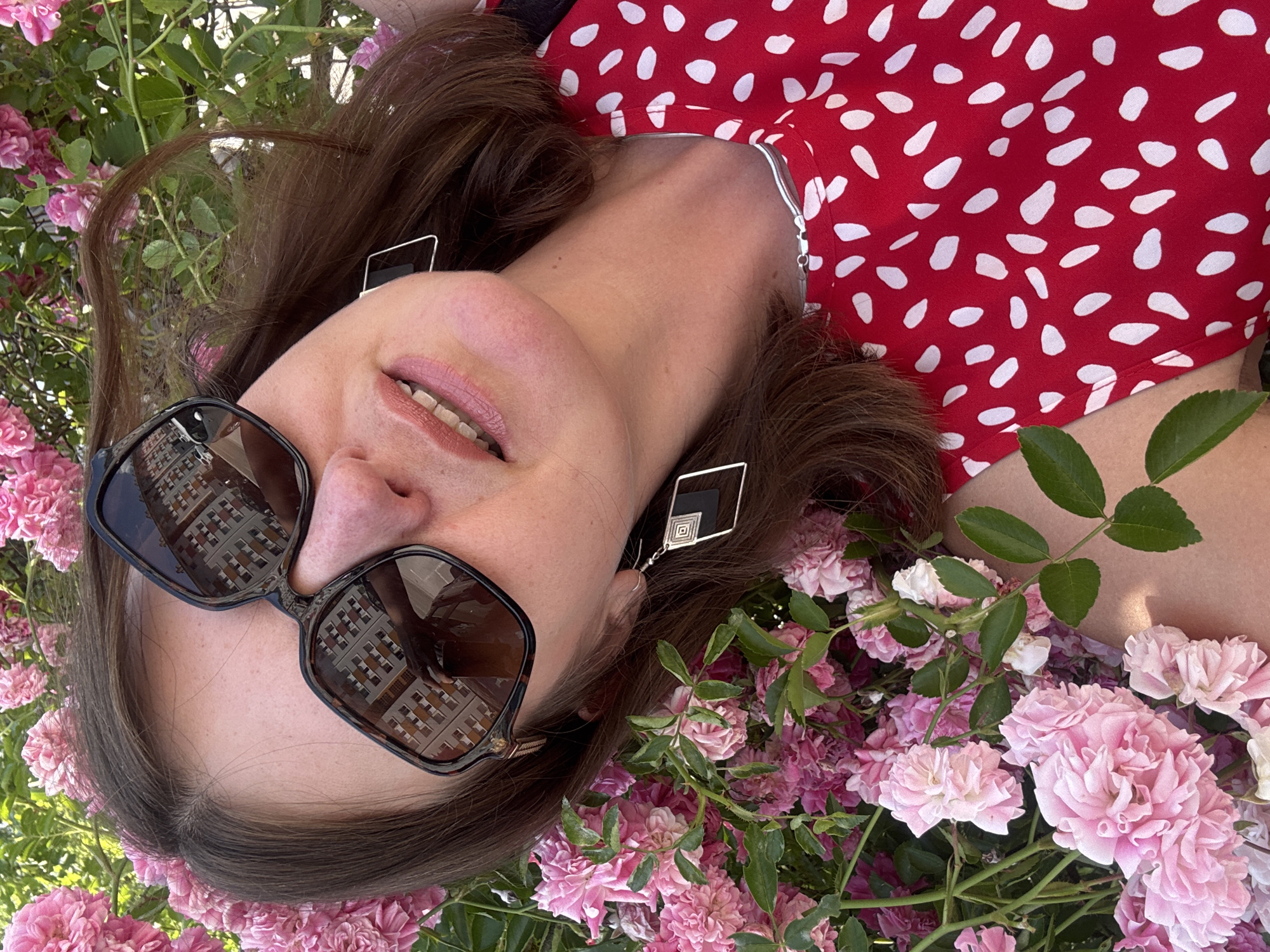 A woman is lying down with her head resting on pink roses, wearing large sunglasses and a red dress with white polka dots. Her face is partially obscured by her sunglasses, but her mouth is open in a smile. She is wearing square earrings with a black inlay. The background is filled with pink roses and green foliage. The image appears to be taken outdoors on a sunny day.