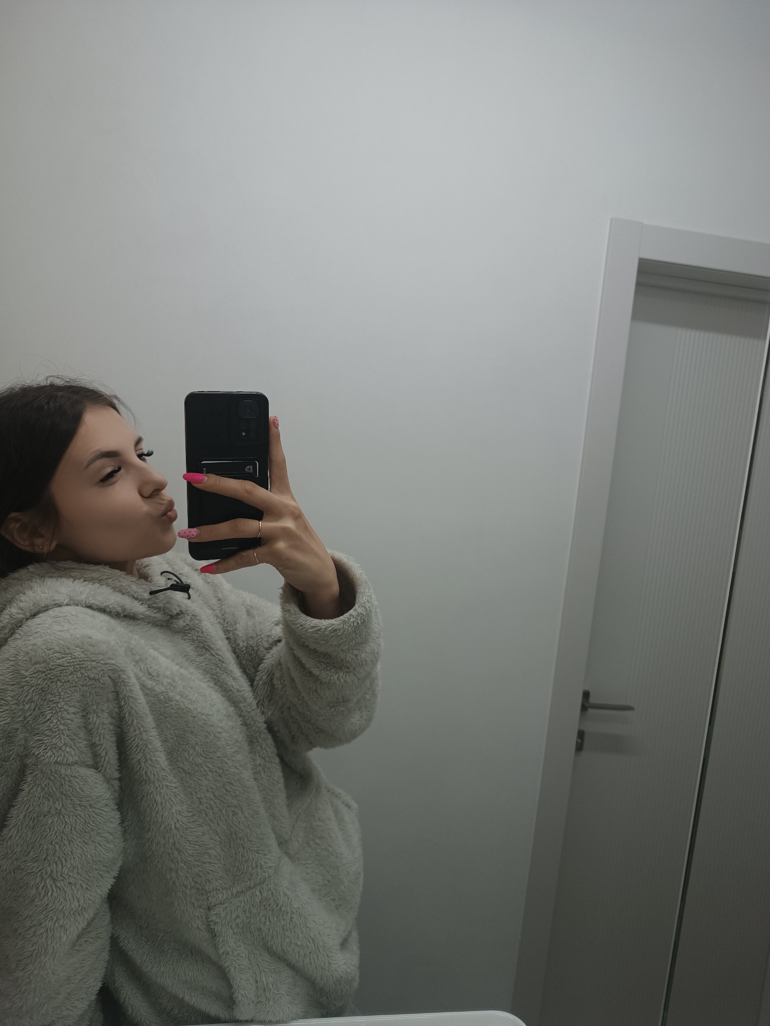 A young woman stands in a bathroom or similar room with white walls and a white door, taking a selfie with a black smartphone. She is wearing a fluffy, light gray hooded jacket. Her fingernails are painted pink, and she is wearing rings on her fingers. She is making a kissing face towards the camera. The background is a plain white wall and a closed white door with a silver handle. There is no indication of Сепыч, Russia.