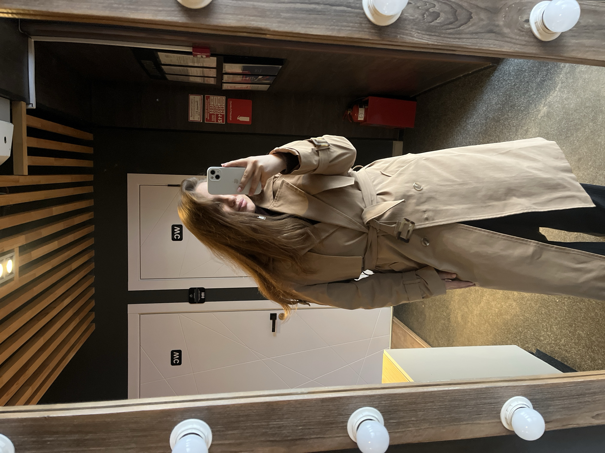 This image is a mirror selfie taken indoors, appearing to be sideways, with the subject holding a white smartphone to capture her reflection. The subject is a person with long, brown hair, wearing a classic beige trench coat over dark attire. The coat is belted at the waist, showing its structured design and a button detail near the collar.
The mirror she is using has a rustic wooden frame adorned with several exposed, globe-shaped vanity lights. Reflected in the mirror is a modern interior space, likely a hallway or common area in a public building in Chaikovsky, Russia. To the left, a wall features decorative wooden slat paneling. To the right, two white doors are visible, each with a black sign displaying the letters "MC," commonly indicating restrooms. Between the doors, there's a black strip with a silver or metallic fixture.
Further in the background, reflected above the doors, is a dark wall adorned with several informational or warning signs. Visible text includes Cyrillic characters. One red sign clearly states "ПОЖАРНЫЙ КРАН" (Fire Hydrant/Valve) and another says "ВНИМАНИЕ!" (Attention!). Below these signs, a red fire extinguisher is mounted on the wall. A light-colored, possibly wooden, rectangular counter or shelf is also reflected near the bottom right of the mirror. The lighting suggests an indoor setting with artificial illumination, and the overall ambiance is neutral, typical for a public space.