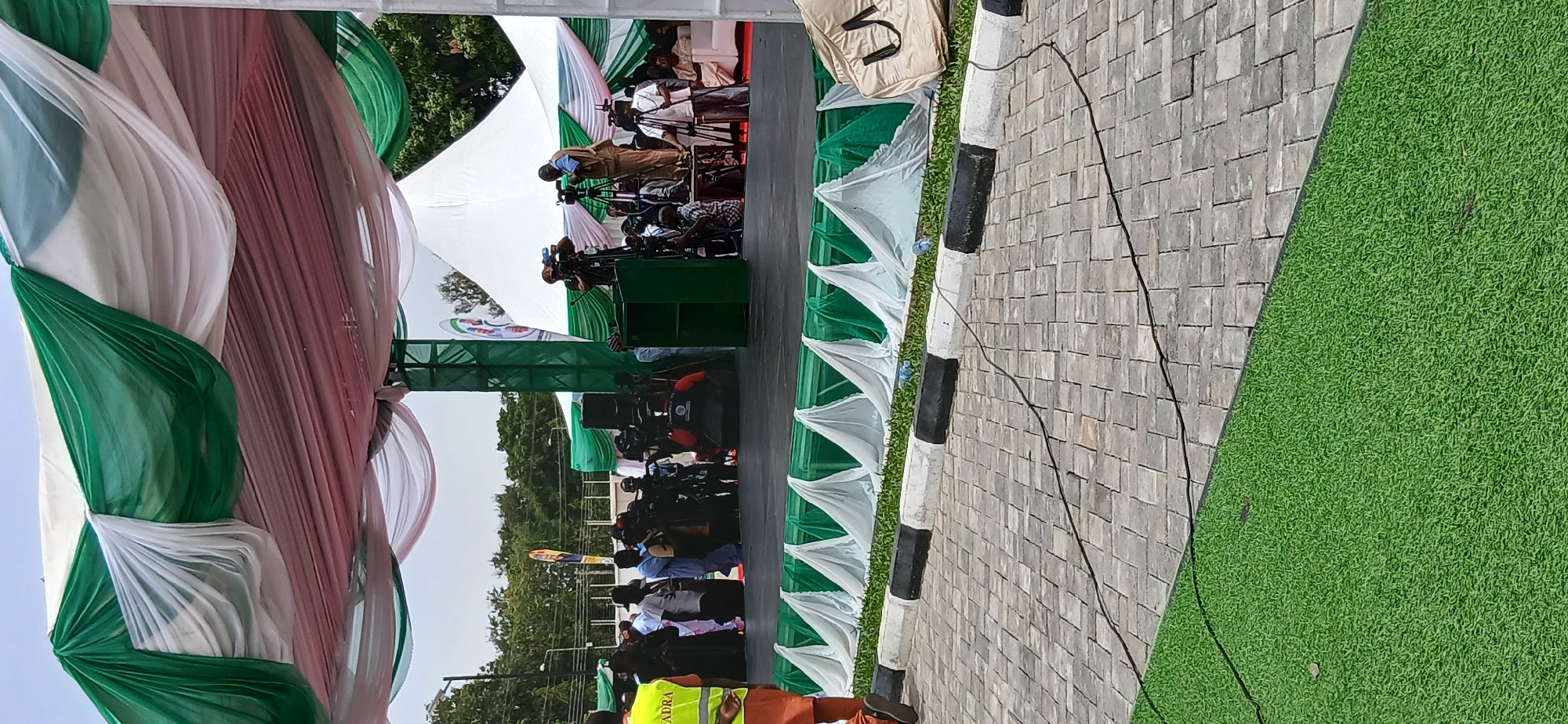 This image captures an outdoor public event, likely a press conference or formal gathering, characterized by a significant media presence. The central focus is on a group of media personnel operating numerous video cameras mounted on tripods, positioned in front of what appears to be a stage or platform.
The event setting includes a large white tent structure, decorated with draped translucent fabric in green, white, and a reddish-brown/pink, creating an elaborate canopy. The stage area features green structural supports and a white backdrop displaying a large circular logo, where the text "NEFRA" is partially visible, along with green and white vertical stripes.
The foreground shows a paved area composed of gray rectangular pavers, bordered by a white curb with distinctive black stripes. Adjacent to this is a strip of vibrant green artificial turf, which extends further to the right. A black cable snakes across the paved surface. Near the curb, two blue plastic bottles are visible.
In the bottom left corner, a person is partially visible, wearing an orange safety vest that displays "ARAN," likely part of a larger word like "RADRA." A beige bag with the letters "VS" clearly printed on it rests near the curb on the right side.
The event is held outdoors during daylight hours under a clear sky, with trees visible in the distant background. The prominent use of green and white in the decorations strongly suggests a connection to Nigeria, whose national flag features these colors. The atmosphere appears orderly and professional, indicative of an official proceeding.