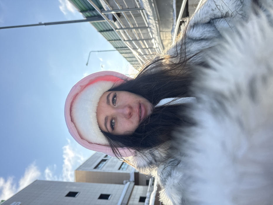 The image presents a close-up, low-angle outdoor portrait of a young adult female in Shul'gino, Russia, taken on a clear, sunny winter day. The subject has long dark hair and is wearing a pink hood with white faux fur lining, consistent with a winter jacket or coat. A light-colored, possibly grey or white, faux fur collar or trim from her outerwear extends into the immediate foreground, creating a soft blur at the bottom of the frame. She gazes directly at the camera with a neutral expression.
In the background, on the left, several multi-story, light-colored urban buildings are visible, showing windows and architectural details. Snow is present on ledges and ground surfaces. On the right, an elevated structure, likely a pedestrian walkway or overpass, features a green sound barrier wall and metal railings, also partially covered in snow. A slender street light pole stands against the bright blue sky, which contains scattered white clouds. The scene is brightly lit by natural sunlight, casting highlights on the snow and structures, and creating a soft glow around the subject.
