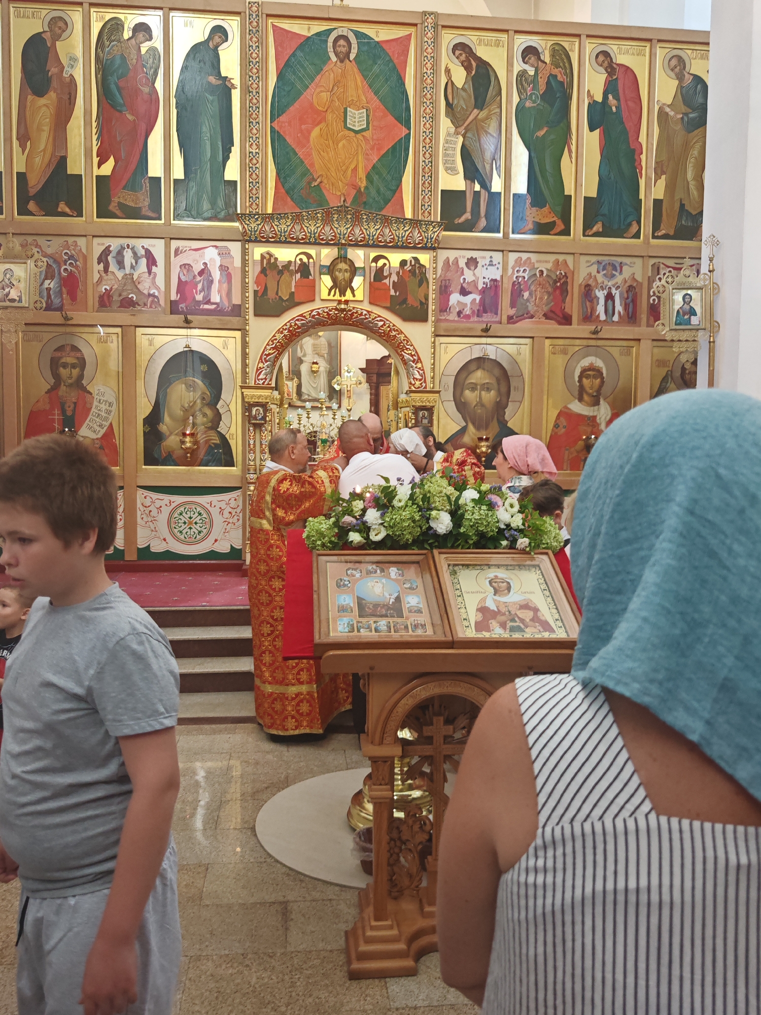 The photograph captures an interior view of an Orthodox church in Varvarovka, Russia, featuring an iconostasis adorned with religious artwork. In the foreground, a young boy in a gray t-shirt and shorts stands to the left, partially obscuring a woman in a blue headscarf and a striped dress, who is facing away from the camera and looking towards the altar area.
The central focus of the image is the altar area, where a priest, dressed in a red and gold liturgical robe, stands facing a group of people. He appears to be engaged in a religious ceremony, possibly administering a sacrament, as he holds a book and gestures with his hands. Other individuals, including men and women, are gathered around him, some wearing headscarves. In front of the altar is a wooden stand displaying religious icons and a floral arrangement.
The background is dominated by the iconostasis, a wall of icons separating the nave from the sanctuary. These icons depict various religious figures, including Jesus Christ, the Virgin Mary, and saints, rendered in a traditional Byzantine style with gold leaf accents. The church interior appears to be richly decorated, with ornate architectural elements and religious imagery consistent with Orthodox Christian traditions. The lighting suggests it is daytime, with artificial light supplementing the natural light entering the space. The floor is tiled, and steps are visible leading up to the altar area.