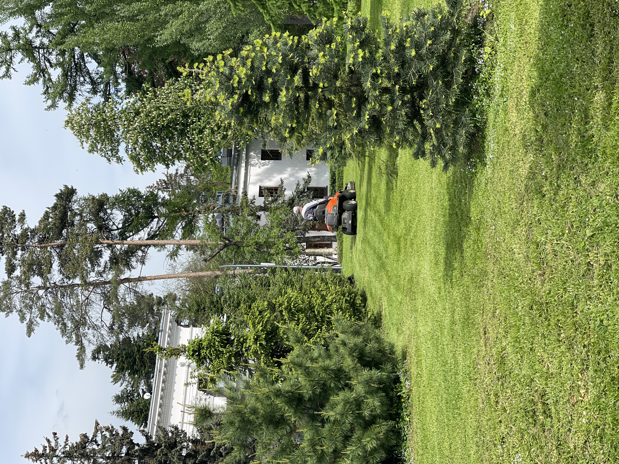 A person riding a lawn mower is seen mowing a lawn next to trees. A building is visible in the background, partially obscured by trees. The scene appears to be in an outdoor setting with a well-maintained lawn. The location is in the city of Unknown, Unknown.