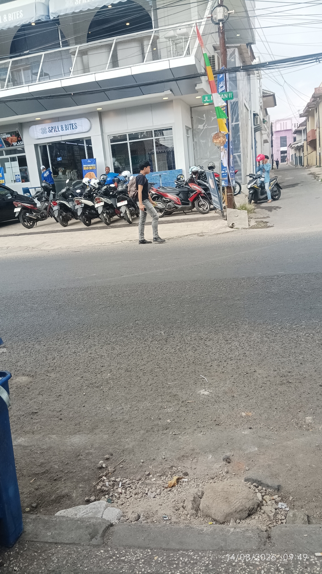 The image captures a street scene in Tasikmalaya, Indonesia, likely in a commercial area. In the foreground, a man in light grey pants and a black t-shirt walks across the asphalt road. He appears to be walking away from a row of parked motorcycles, most of which are scooters. Behind the motorcycles is a building with the prominent sign "SPILL & BITES" above glass storefronts. The building has multiple levels with reflective windows and grey detailing. To the right of the building, a street sign reads "JL. MAN II," indicating the street name. Further down the street, more buildings and a few more motorcycles are visible. A person in blue is seen inside the "SPILL & BITES" establishment. Another person in a blue jacket is on a motorcycle in the background. The environment suggests a typical urban setting with a mix of commercial activity and transportation infrastructure. The timestamp in the bottom right corner indicates the photo was taken on 14/08/2023 at 09:49.