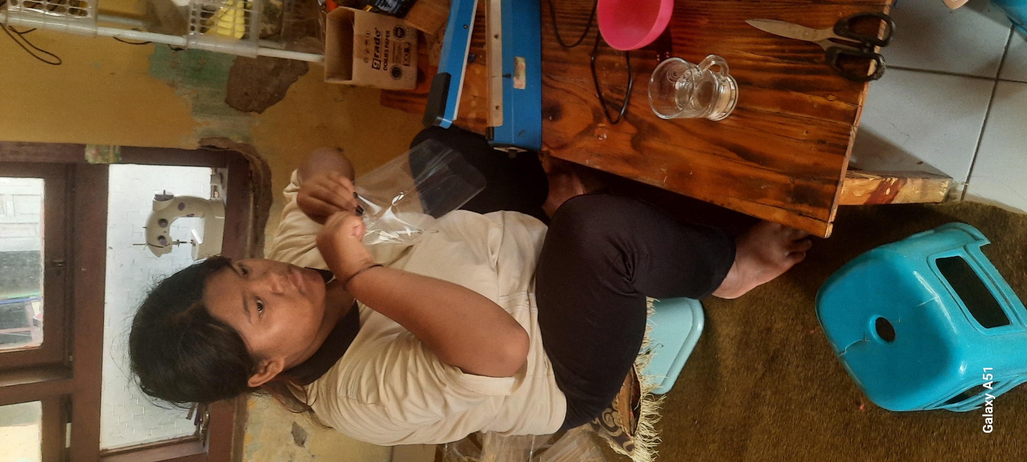 In Ploso, Indonesia, a person is seated on a light blue stool in what appears to be a workshop or home environment. The individual is wearing a cream-colored t-shirt with a dark collar and dark pants, and is in the process of sealing a clear plastic bag. Their right hand holds the top of the bag open, while their left hand appears to be operating a heat-sealing machine. The machine, a blue device with black handles, is positioned on a wooden table.
The table is cluttered with various items, including a small glass mug filled with liquid, a pink bowl, a pair of scissors, and a cardboard box labeled "GRADE DOLBIES PAPER." The background shows a window with wooden frames and a small sewing machine visible through the glass. The walls appear to be aged and possibly damaged, with patches of peeling paint. A white plastic shelf unit is also visible in the background, on the right side of the image. The floor is covered with a woven mat and tiles. The overall scene suggests a workspace where items are being packaged.